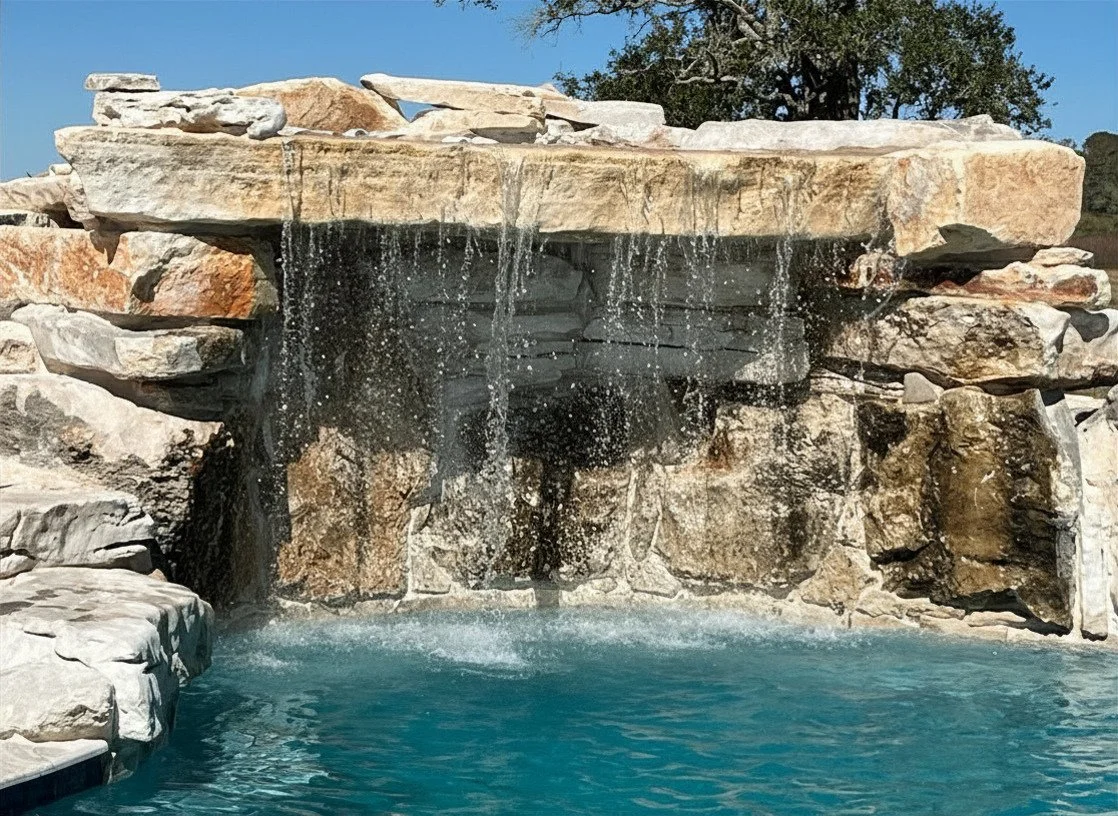 A small outdoor pool with a rocky waterfall feature, rocks above and around the waterfall, cascading water, and a clear blue sky with trees in the background.