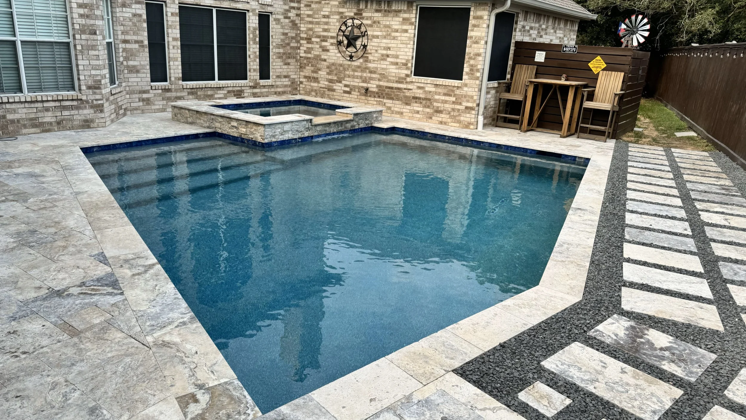Residential backyard with a swimming pool and hot tub, brick house exterior, decorative star wall art, outdoor furniture, windmill, and a wooden fence.