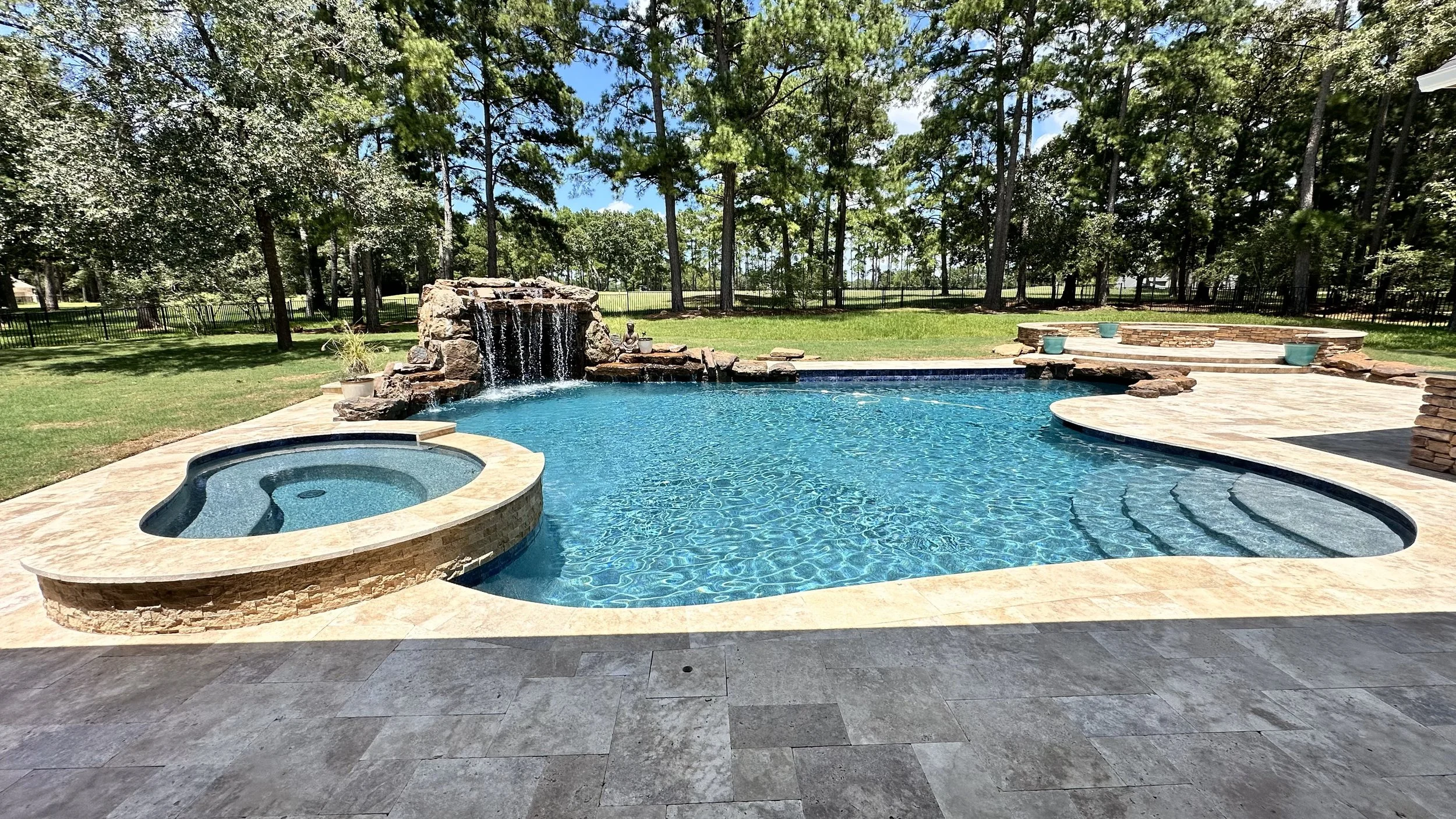  backyard swimming pool with waterfall feature, built-in hot tub, and stone deck, surrounded by tall trees and a fence in the background.