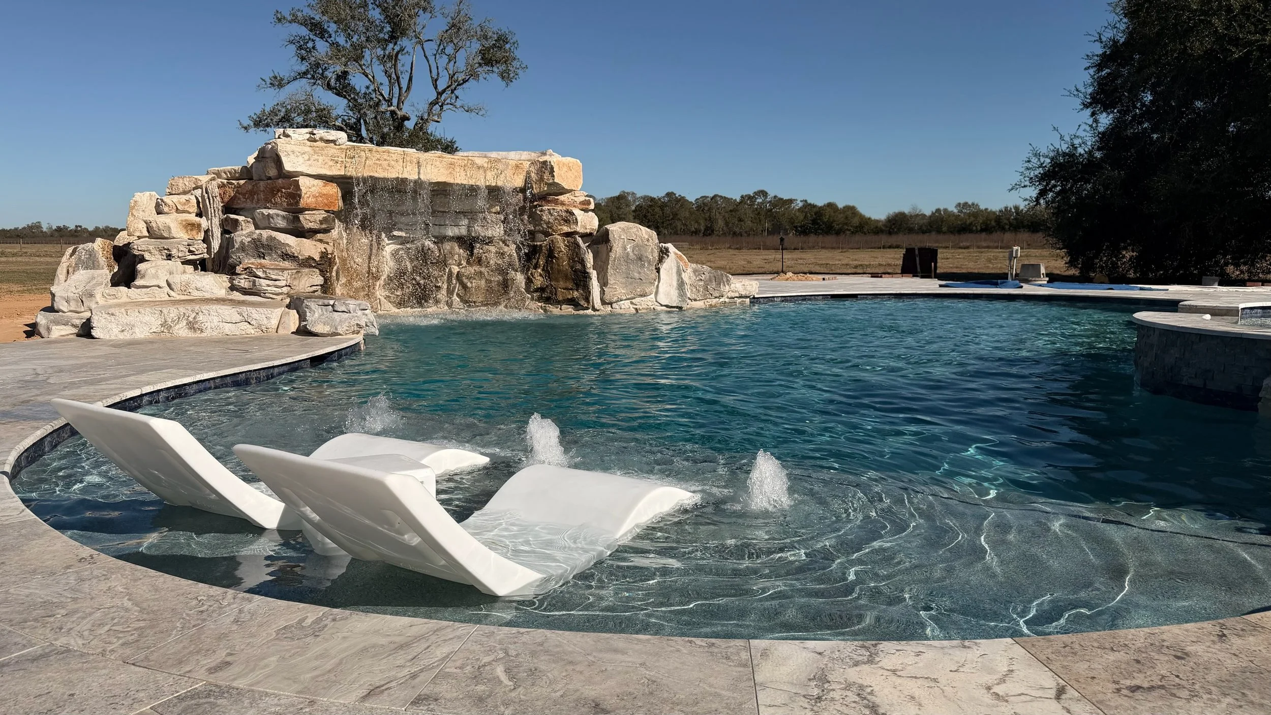 A backyard swimming pool with a rock waterfall feature, two white lounge chairs partially submerged, and an open sky with trees in the background.
