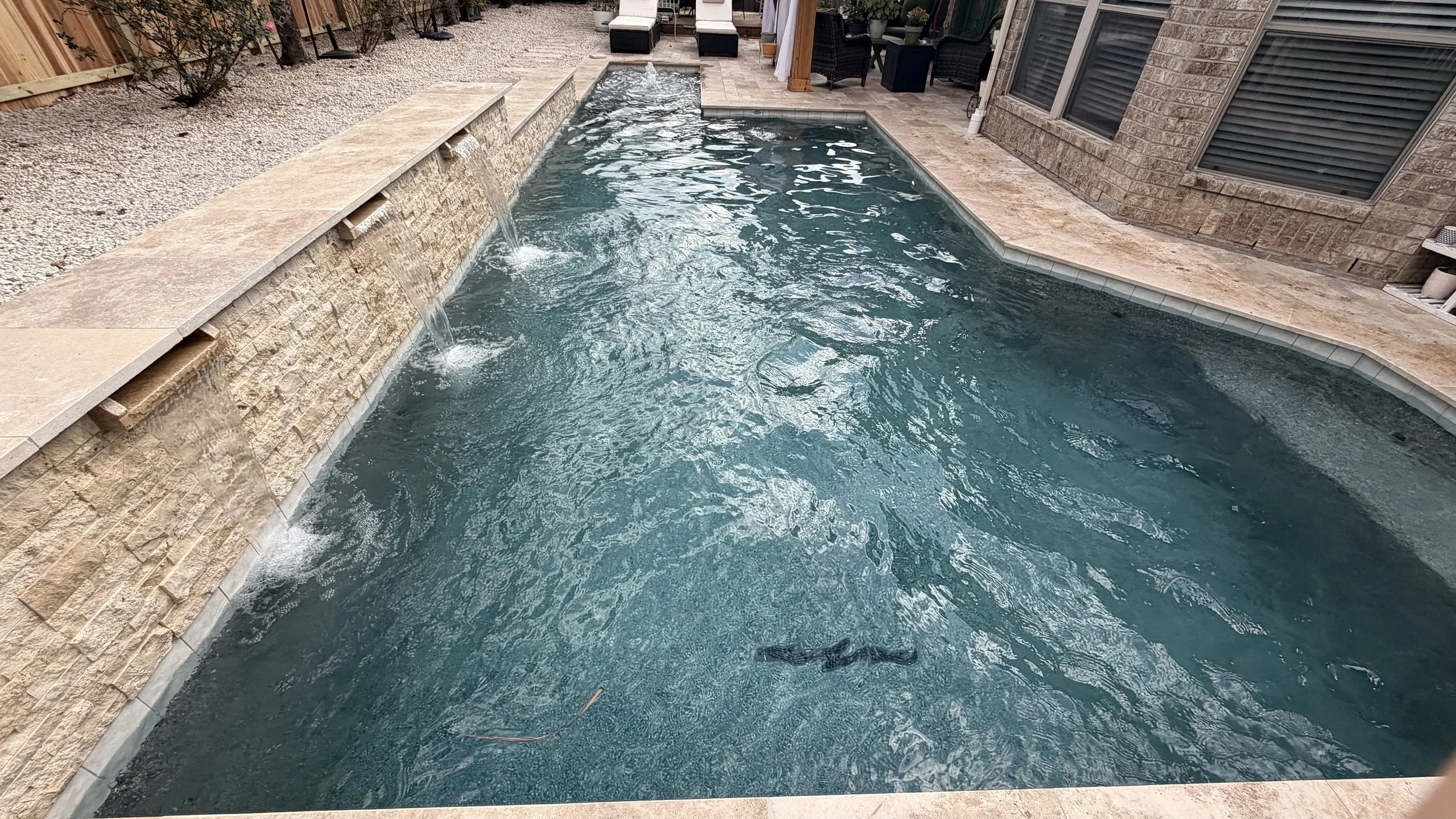 Backyard pool with stone and tile edging, small water fountain, and outdoor seating area with chairs and table. Brick house wall with windows in the background.