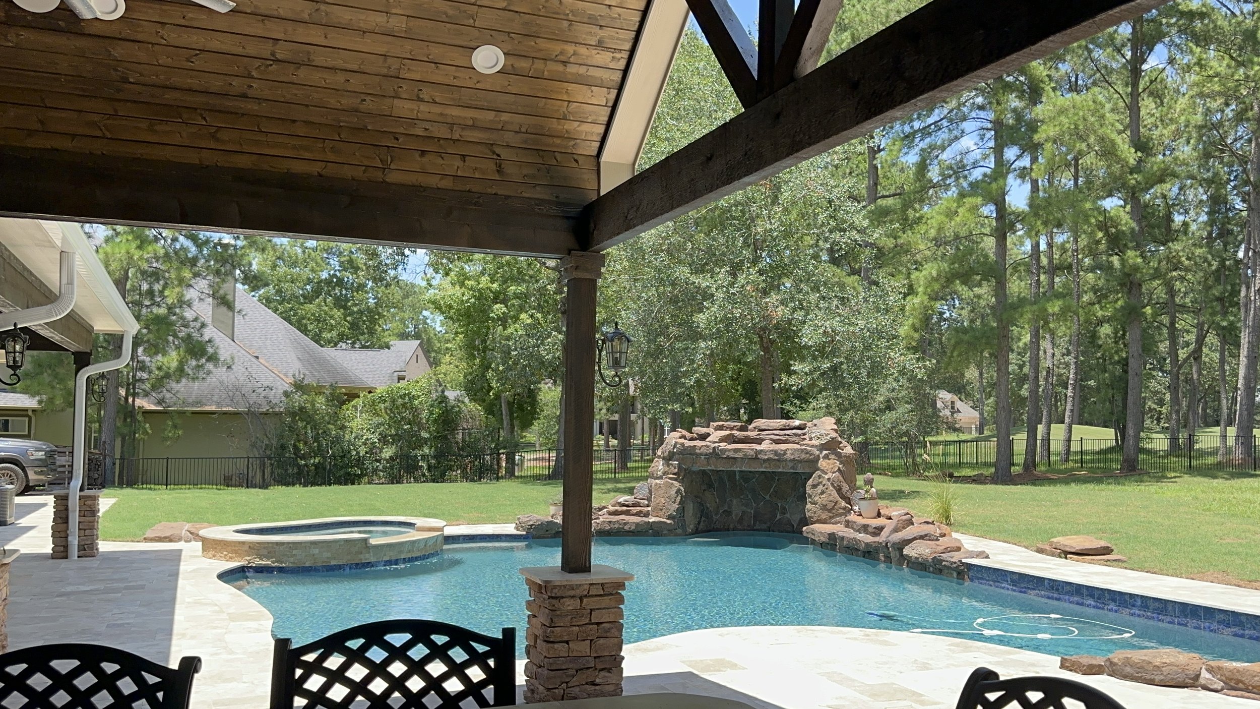 Backyard view of a swimming pool with a rock waterfall feature, outdoor furniture, and surrounded by trees and neighboring houses.