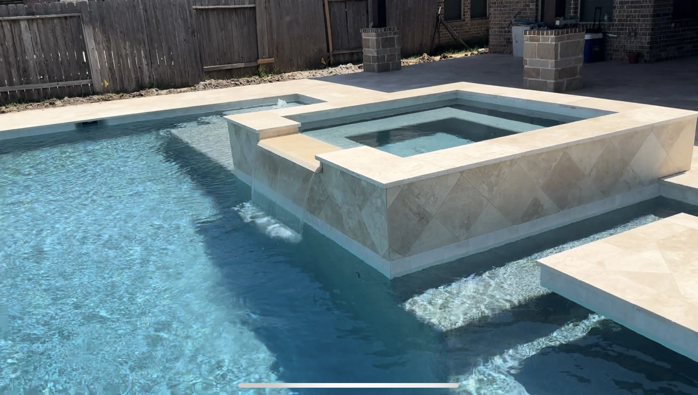 Residential backyard pool with built-in hot tub and water feature, surrounded by a concrete deck and a wooden fence