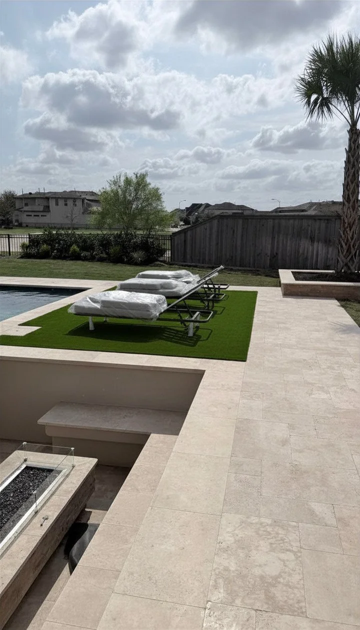 Empty backyard with a row of covered lounge chairs on artificial grass, a swimming pool, a palm tree, and houses in the background under cloudy sky.