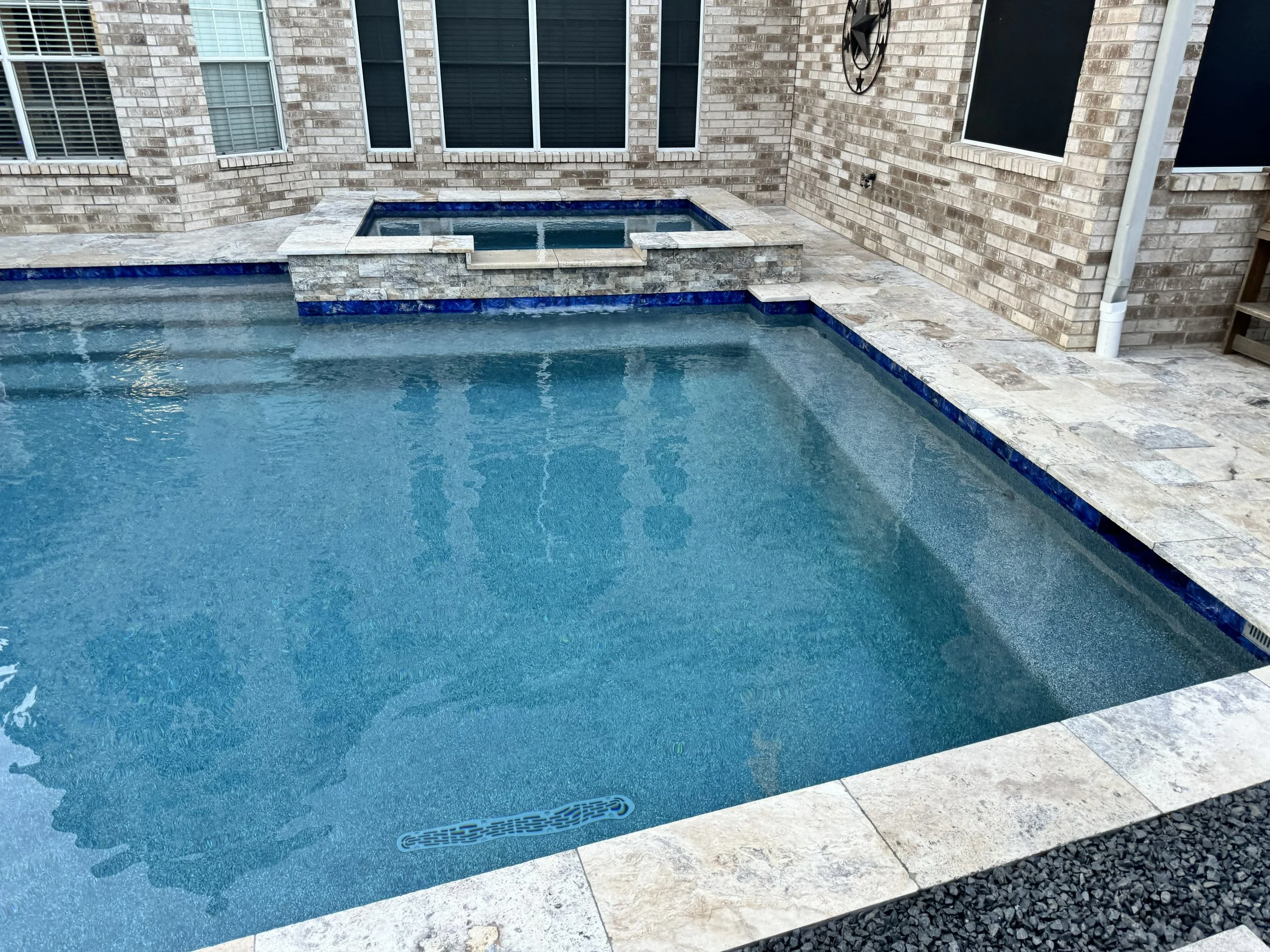 A backyard pool with a built-in hot tub, surrounded by stone tiles, against a brick house wall with multiple windows.
