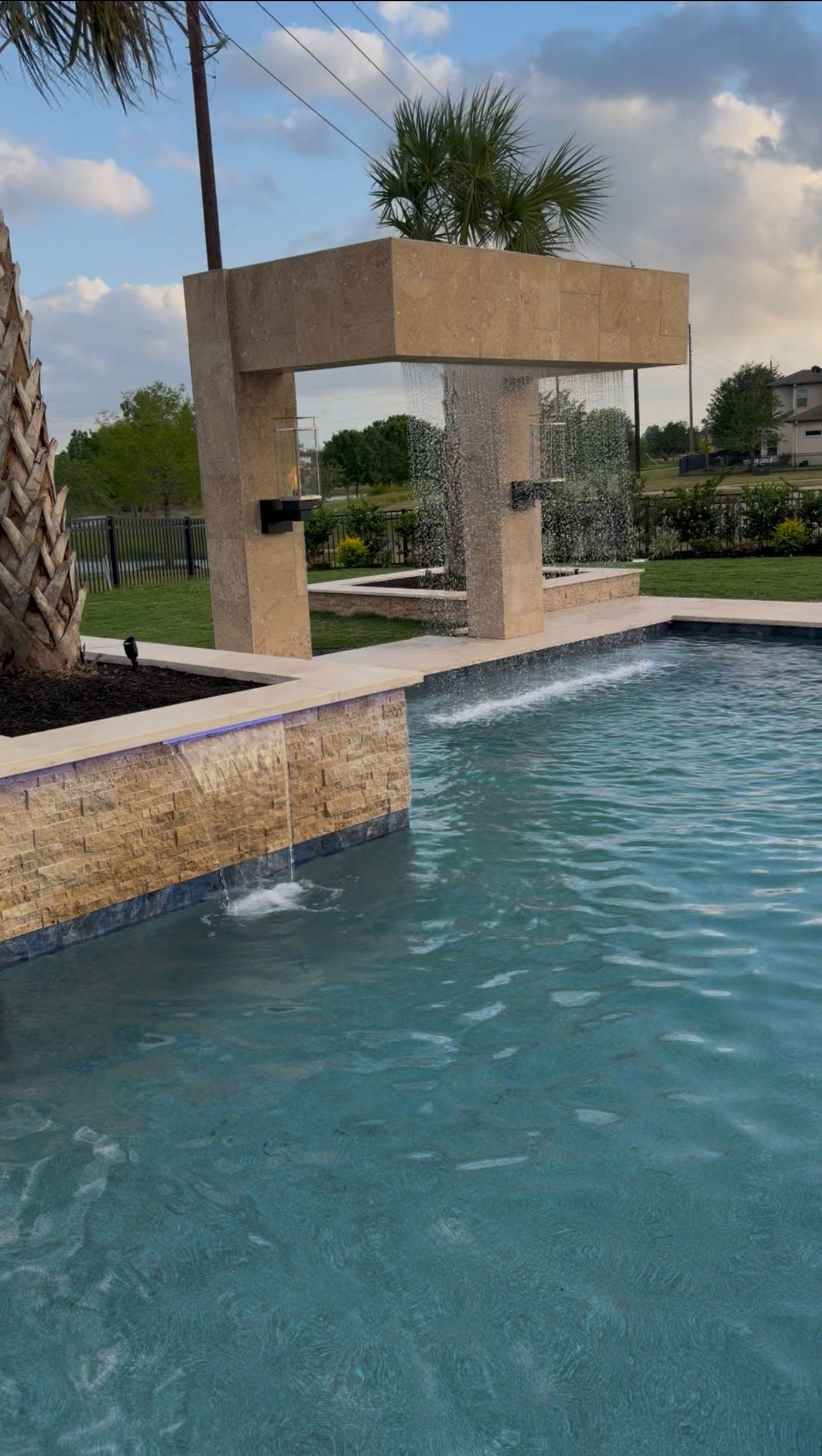 A modern backyard swimming pool with a water feature fountain, a stone waterfall edge, and a beige architectural structure with water cascading down.