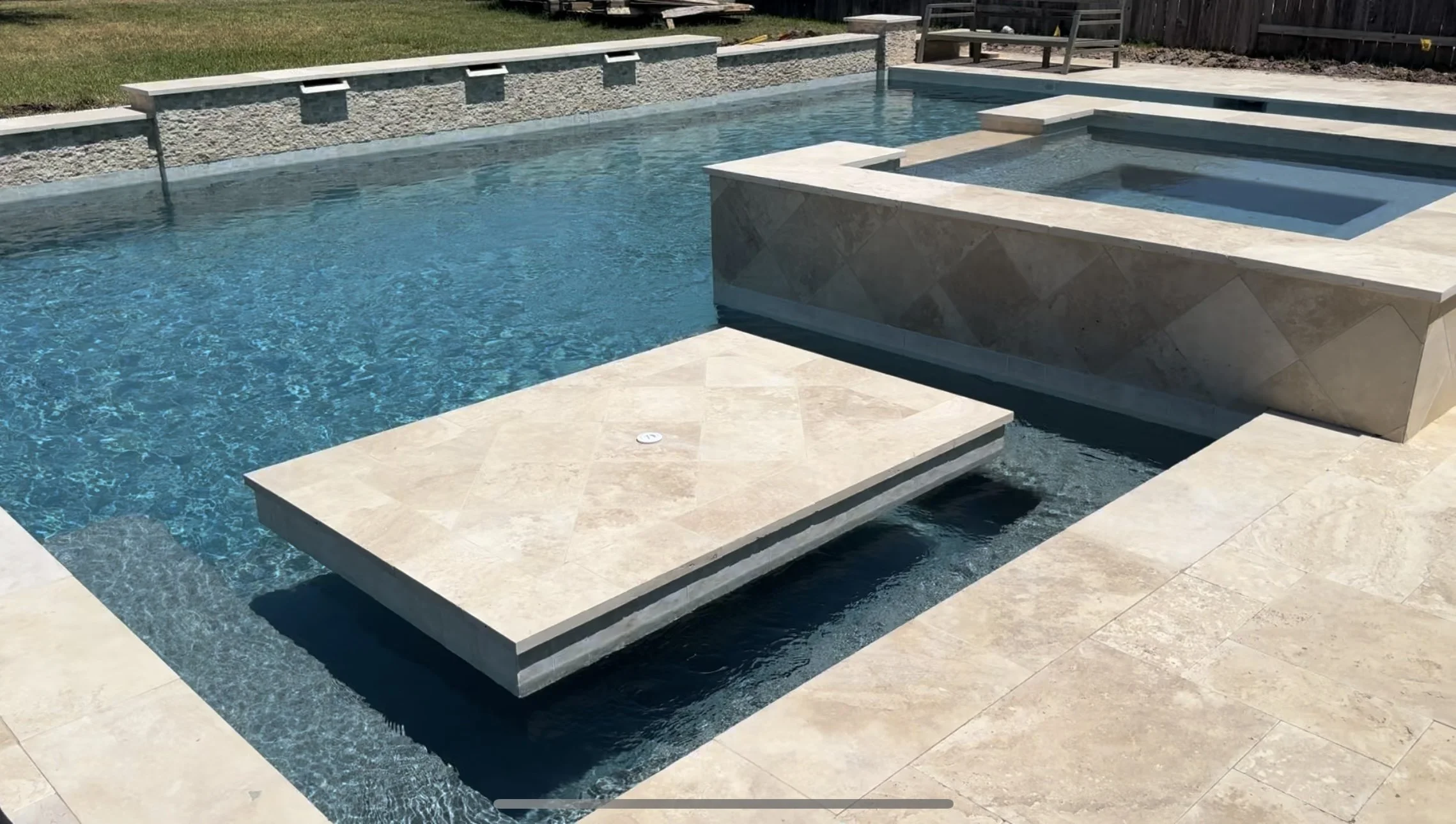 Modern backyard swimming pool with built-in hot tub, surrounded by beige stone paving and a grassy area, fenced yard in the background.