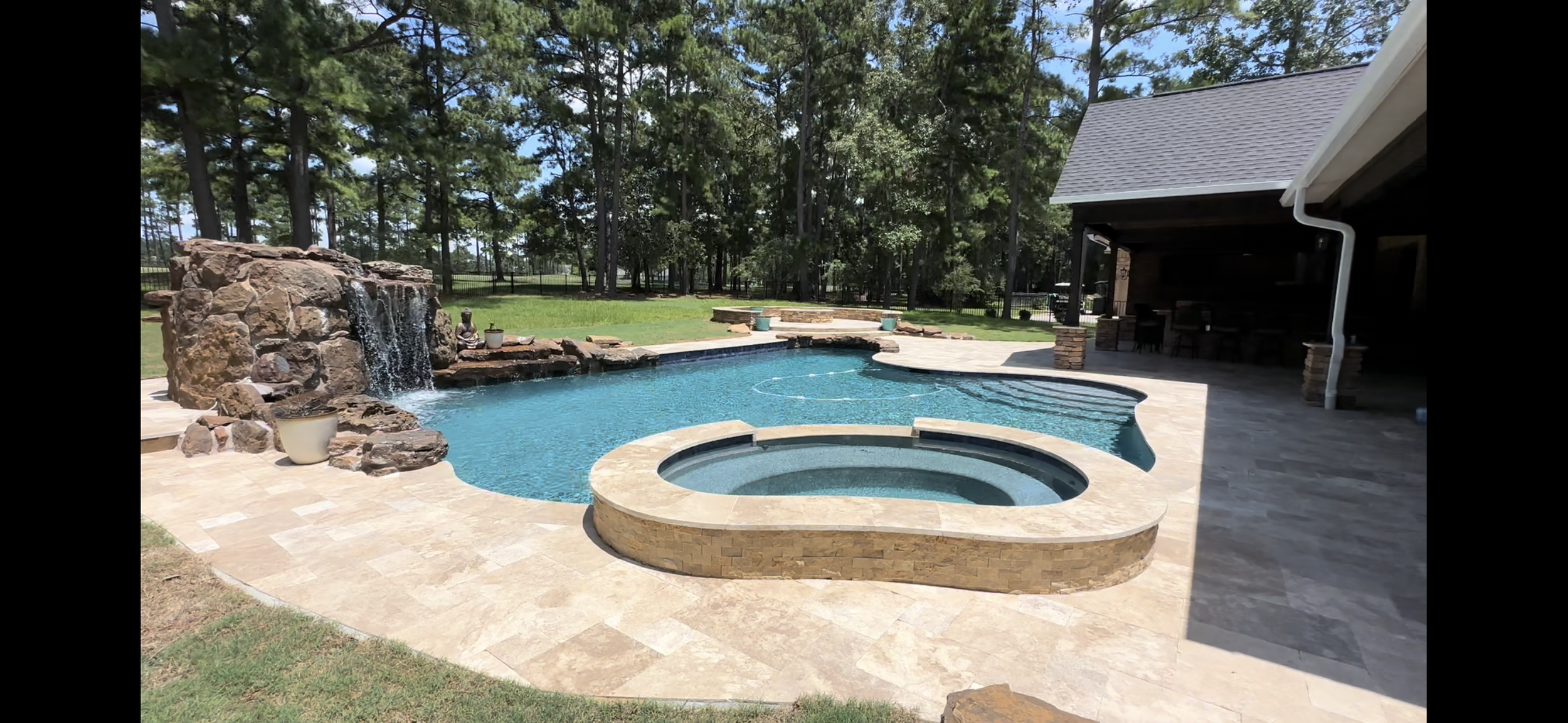 Backyard scene with a swimming pool and hot tub, stone waterfall feature, surrounded by a screened fence and tall trees, with a covered patio area on the right.