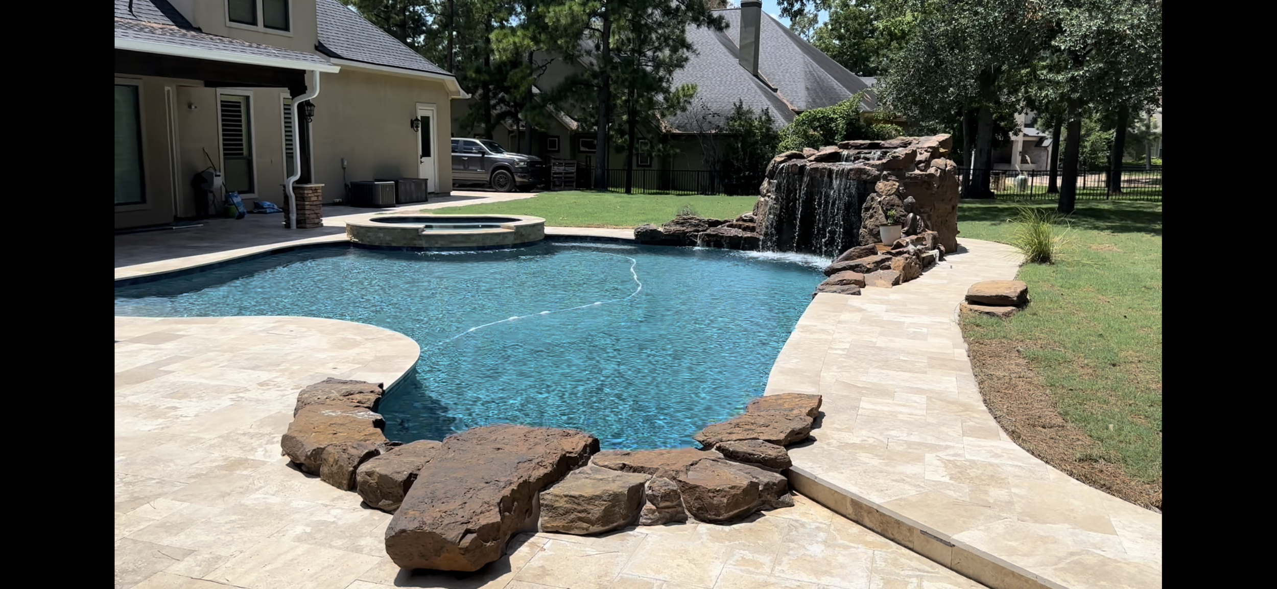 Backyard with a swimming pool, rock waterfall feature, a hot tub, and a stone patio, with residential houses and trees in the background.