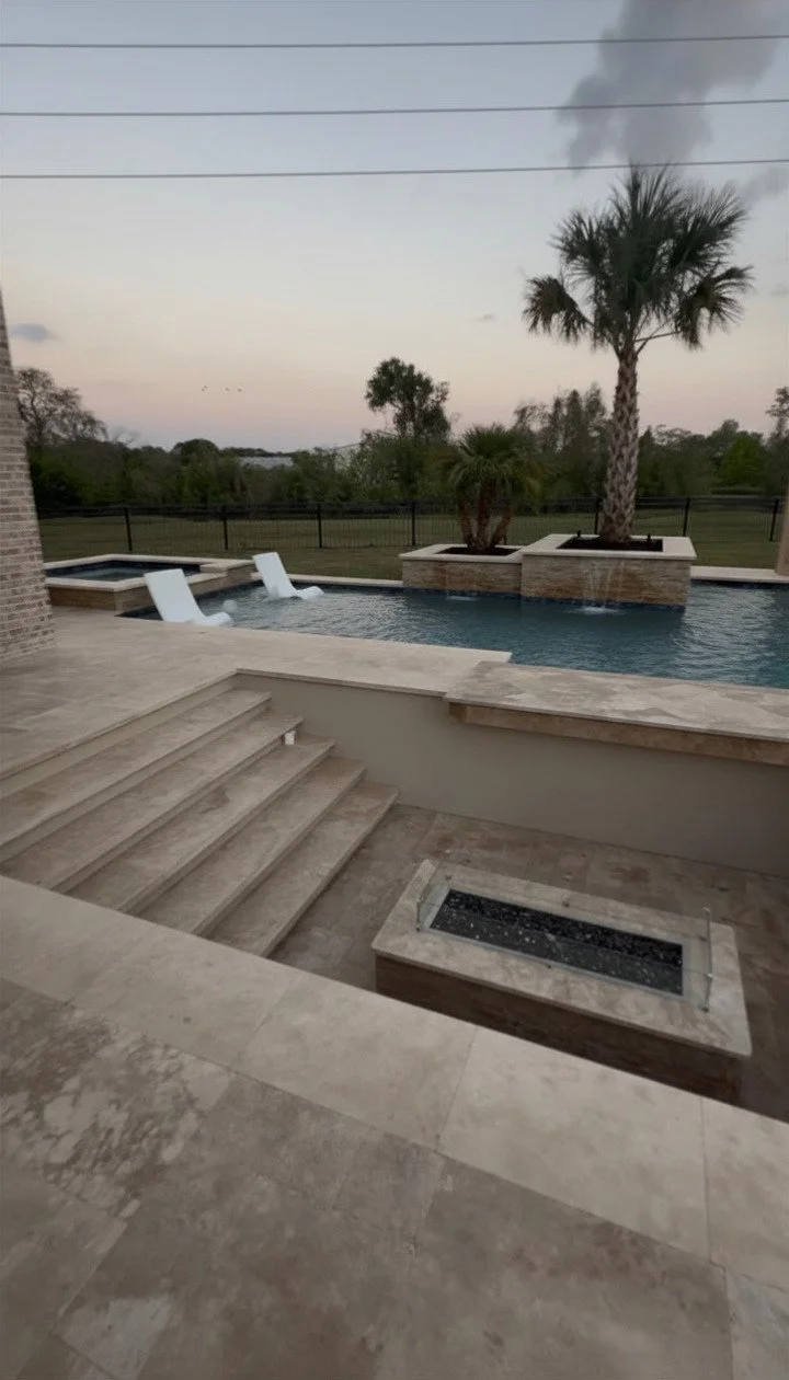A backyard swimming pool with two white lounge chairs on a raised platform, surrounded by palm trees, a grassy yard, and a black fence in the background. The sky is light with a hint of sunset colors.