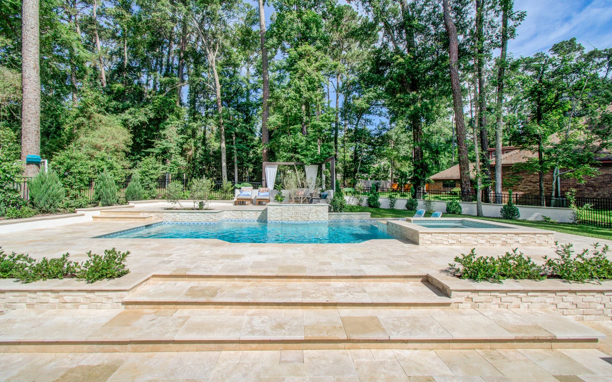 Backyard with in-ground pool surrounded by beige stone paving, with outdoor seating area with white canopy and lush green trees in the background.