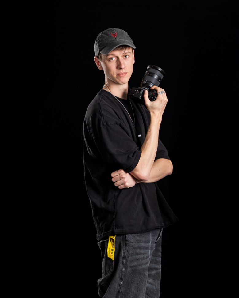 Einer der drei Gründer, Daniel Krieger, mit schwarzer Cap, schwarzem T-Shirt und grauen Jeans hält eine Kamera in der Hand, steht vor schwarzem Hintergrund.