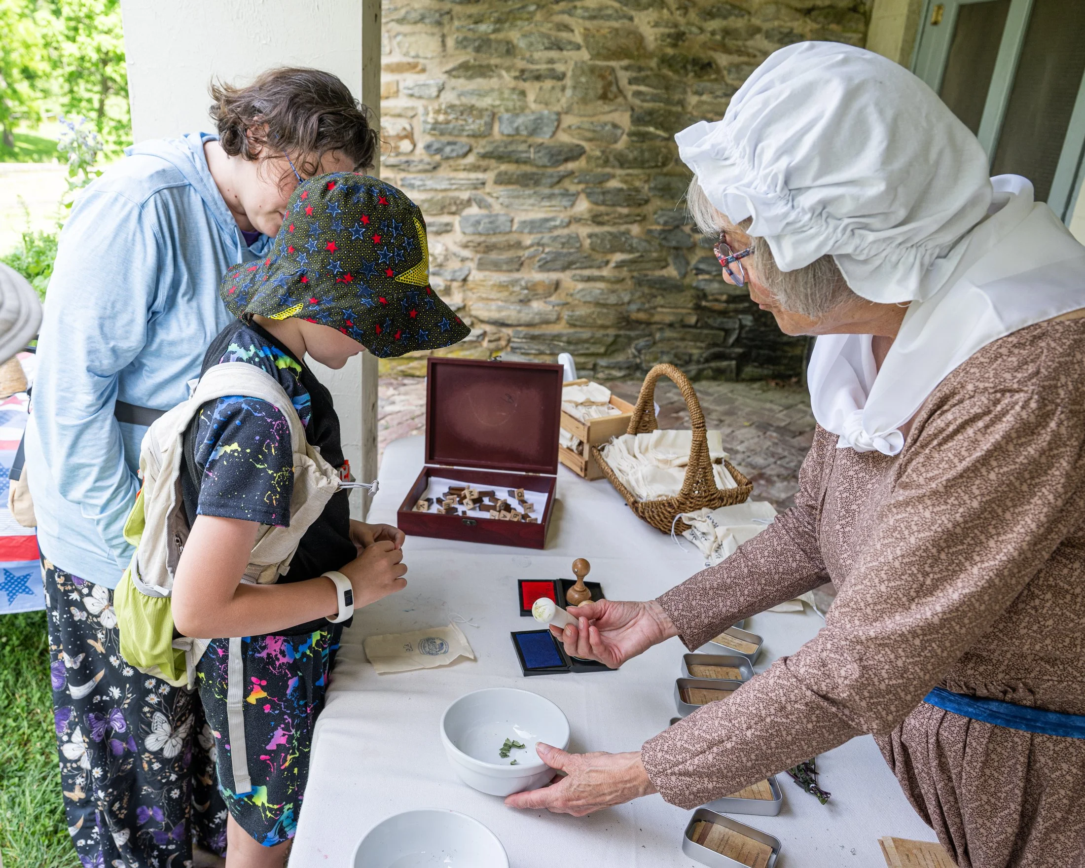 Diane Whitcomb demonstrates crushing herbs in a mortar with Flynn and Tyler Cordes, of Elizabethtown, during Rock Ford America250 Family Day on Sunday, June 7, 2026. Whitcomb taught visitors about the medicines Revolutionary War soldiers carried in t