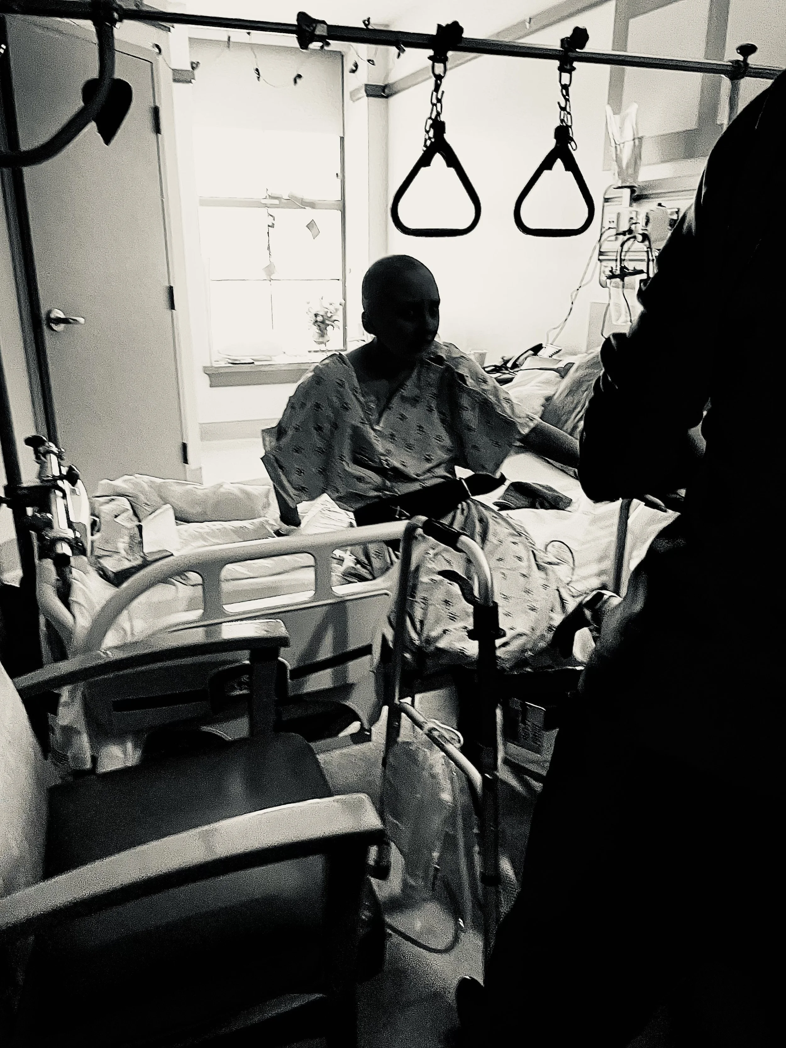 Grayscale photo of Ava in a hospital room seated on a bed, wearing a hospital gown. There is medical equipment such as a walker, IV stand, and overhead trapeze in the room.