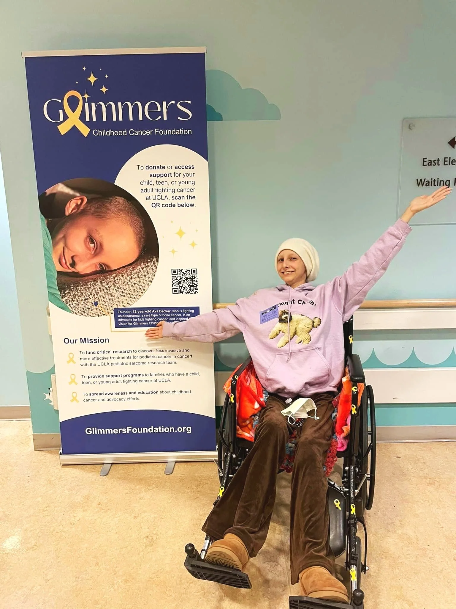 Ava in a wheelchair smiling with arms outstretched, next to a Glimmers Childhood Cancer Foundation banner.