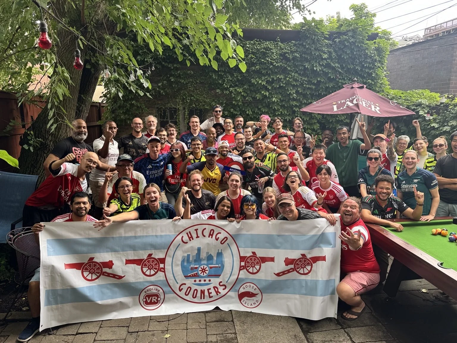 A large group of people, mostly wearing soccer jerseys, gathered outdoors in a backyard with trees and a brick wall in the background. They are smiling, cheering, and holding a large banner that reads "Chicago Cooners." Some are making celebratory gestures, and there is a pool table with billiard balls in the foreground, along with outdoor furniture and a large umbrella.