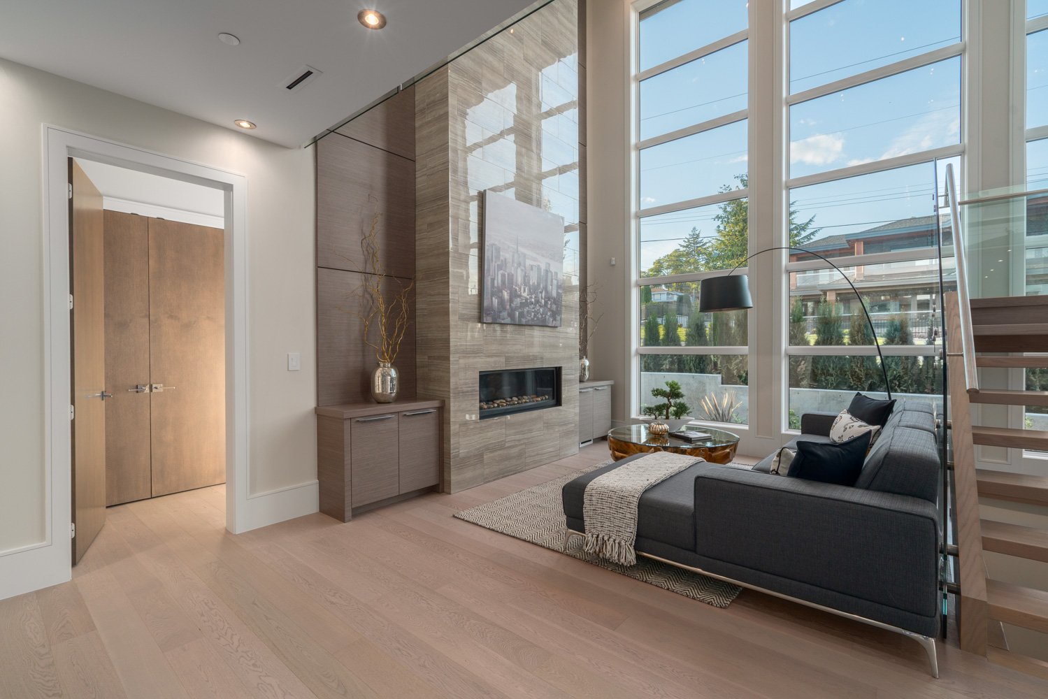Modern living room with large windows, a contemporary fireplace, gray sofa, glass coffee table, and open stairway.