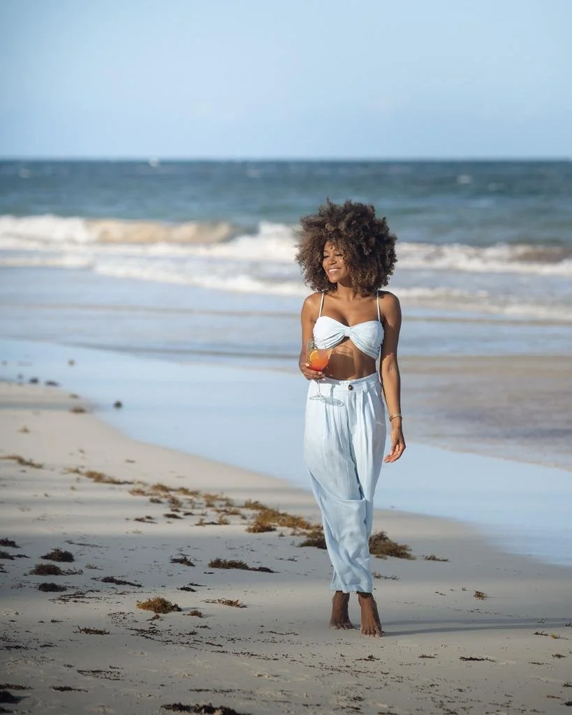 Mujer caminando por la playa con un traje de baño blanco y un vaso de bebida en la mano, con el mar y el cielo azul de fondo.