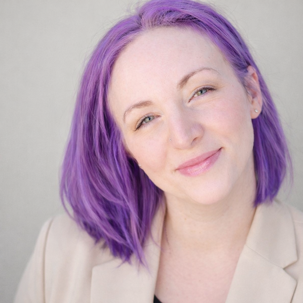 Close-up of a woman with purple hair, light skin, and a subtle smile, wearing a beige blazer.