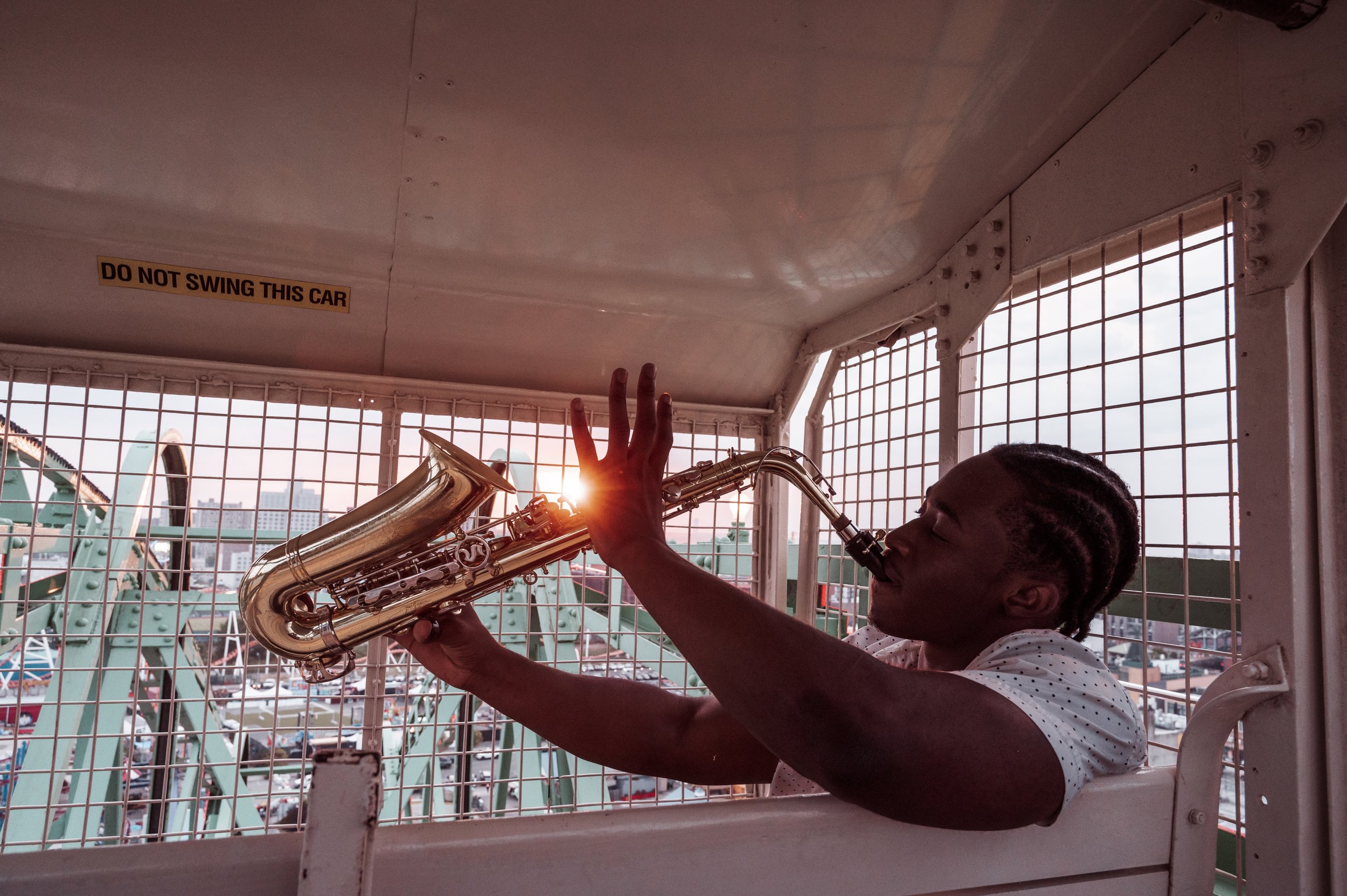 A young man playing a saxophone on a Ferris wheel at sunset, with an outdoor amusement park in the background and a sign that reads 'Do Not Swing This Car' inside the Ferris wheel cabin.