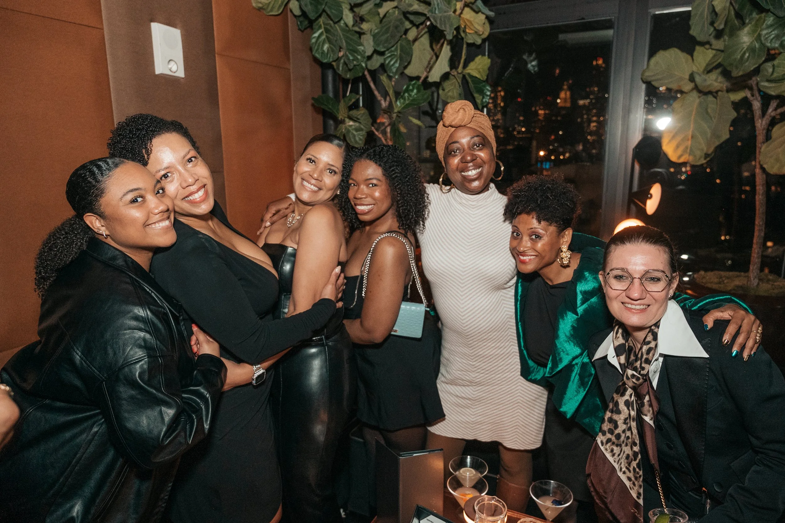 Group of eight women at a party, smiling and posing for a photo indoors with city lights visible through large windows behind them.