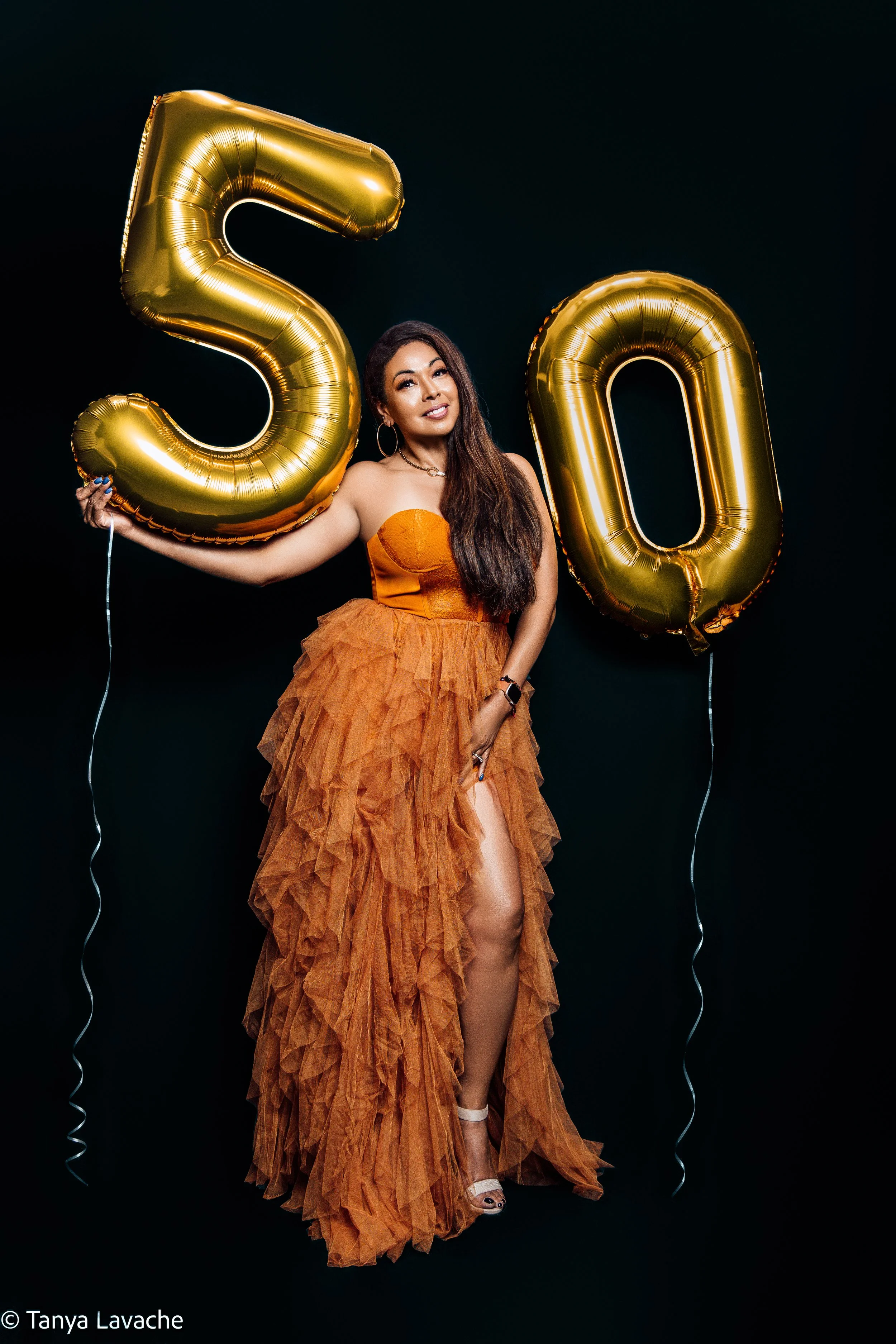 A woman in an orange gown holding gold '50' balloons against a black background.