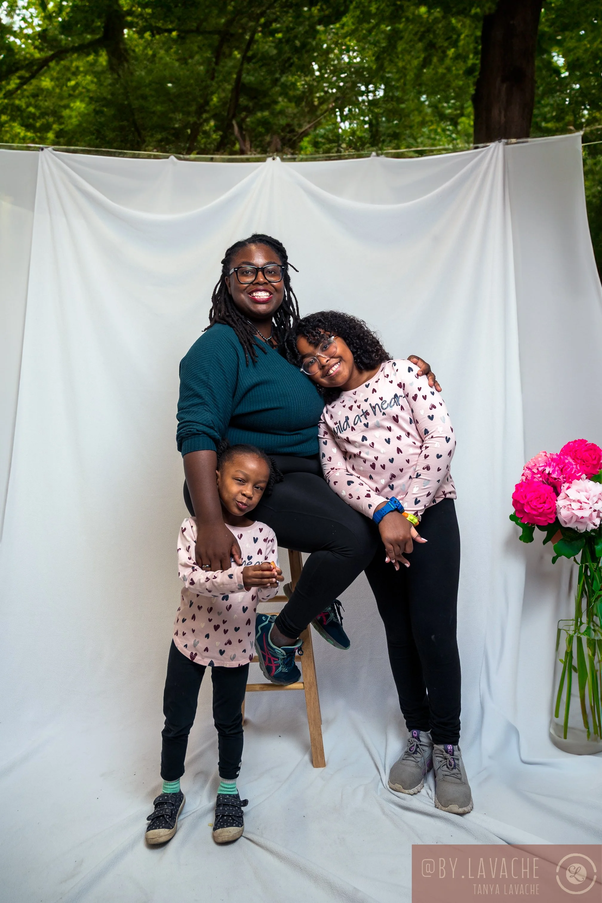 A woman and two young girls posing and smiling in front of a white backdrop outdoors, with trees and green foliage in the background. The woman is sitting on a stool, while the girls are standing beside her. One girl is resting her head on the woman's shoulder, and the other girl is standing in front, holding an object. There are pink flowers in a vase to the right.