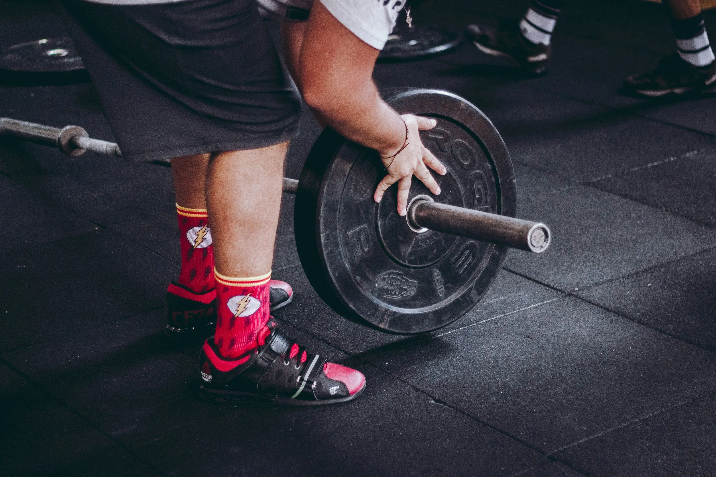 A person wearing red and black sports shoes and red socks with white lightning bolts lifting a barbell in a gym.