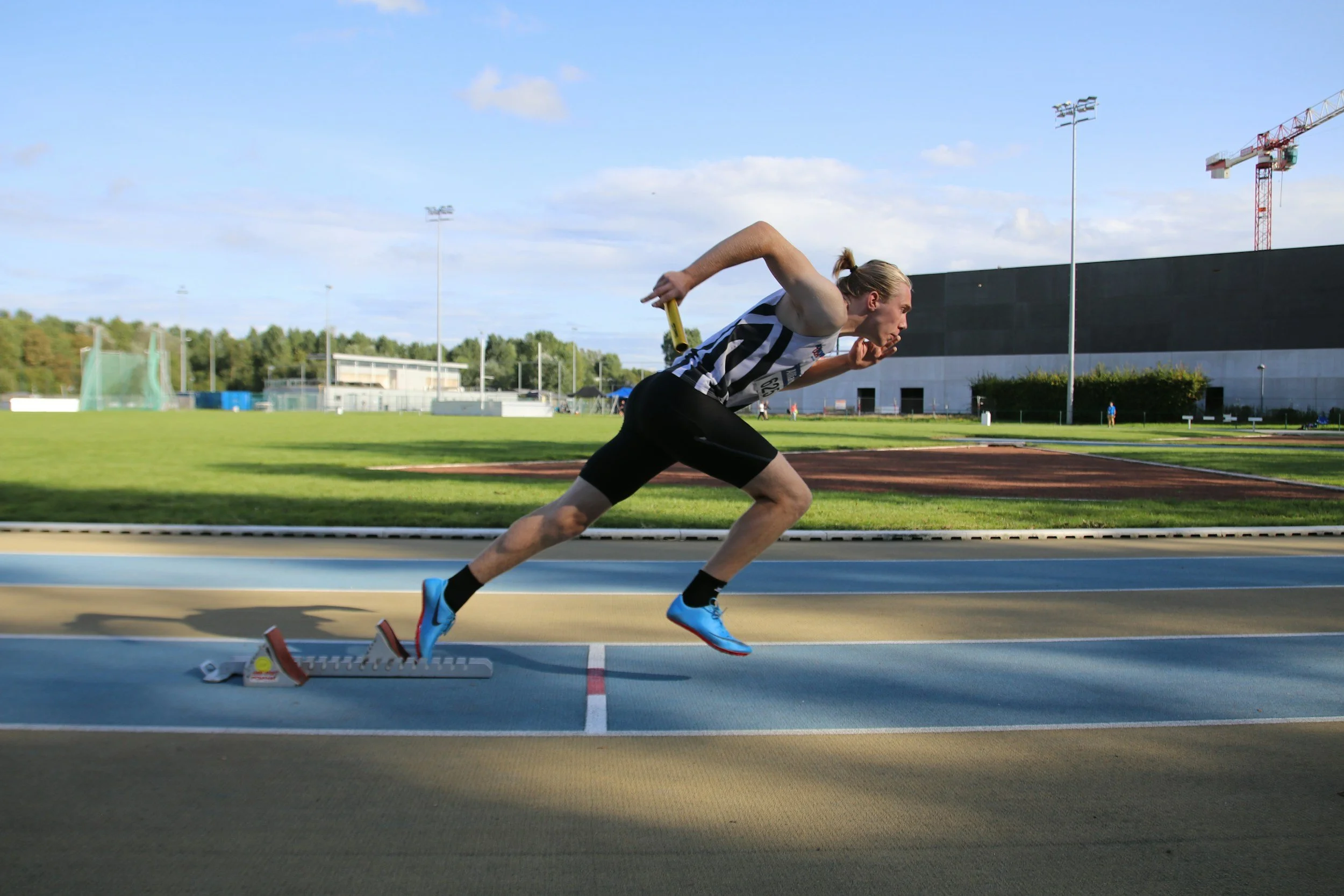 A male athlete in track shorts and a striped jersey is lunging forward in a sprint start position on a running track, with starting blocks in front of him.