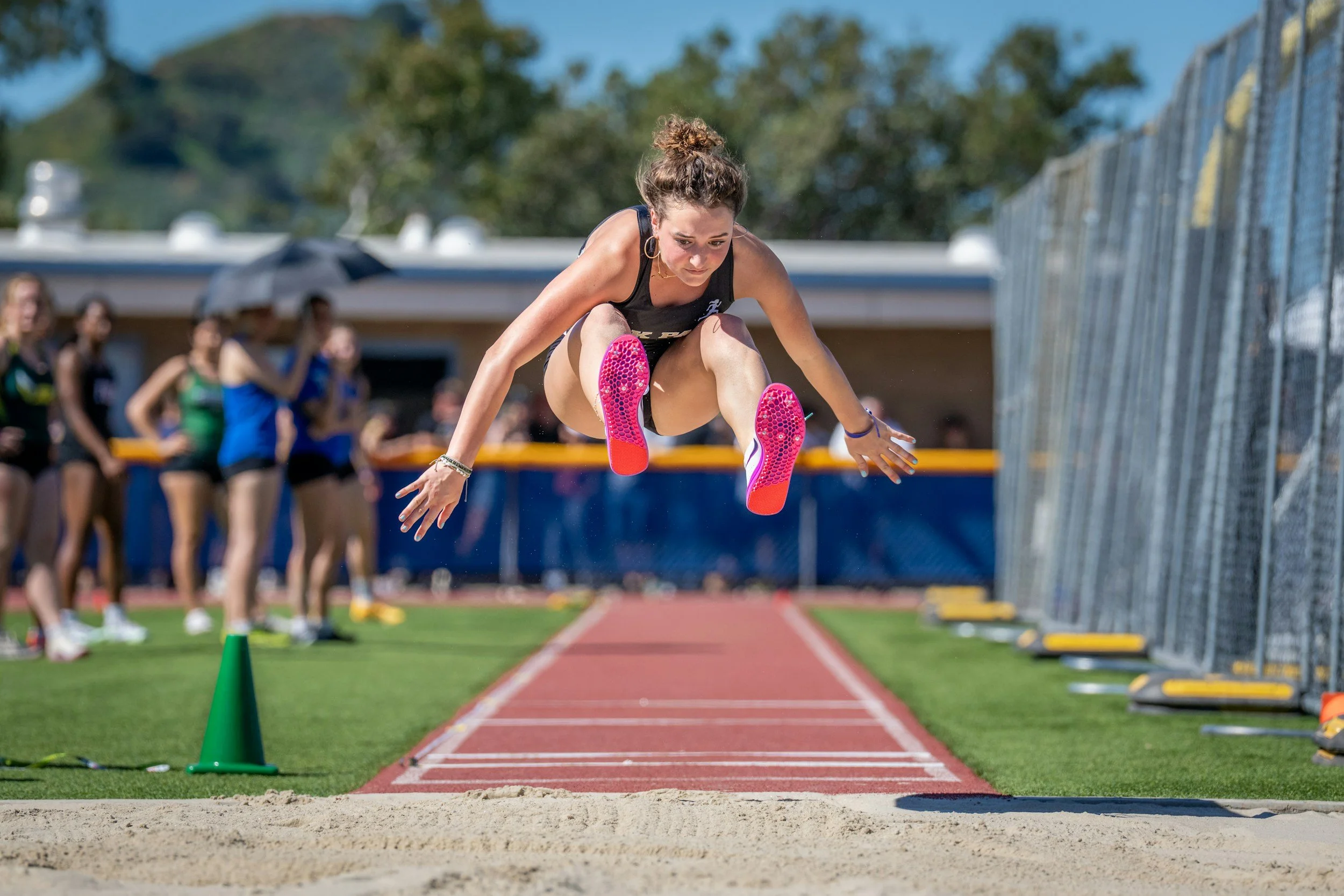 Female athlete mid-air in long jump at outdoor track and field event with spectators and officials in the background.
