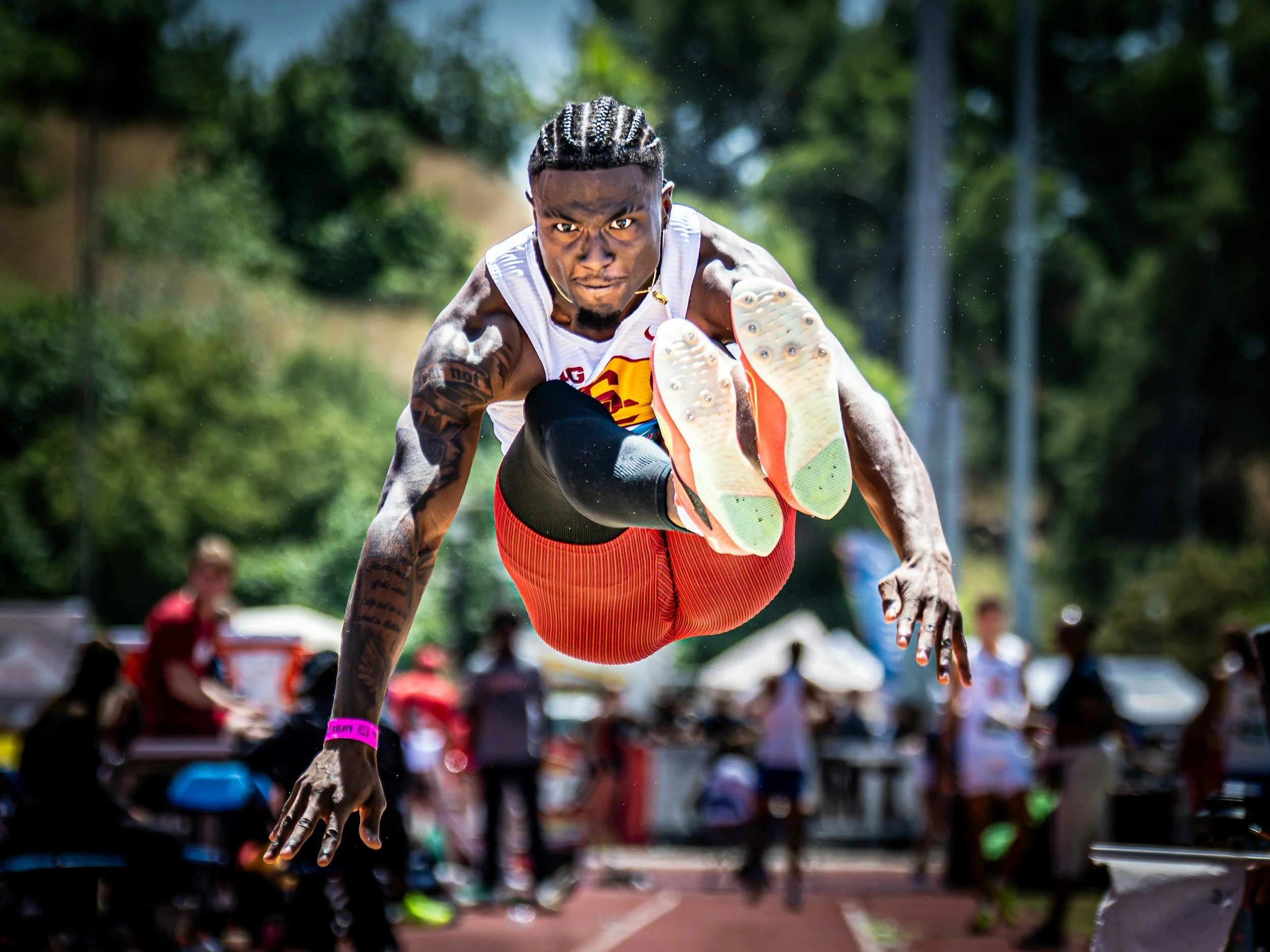 Male athlete with cornrows in a race or sprint, leaning forward with intense focus, outdoors with a blurred crowd and green trees in the background.