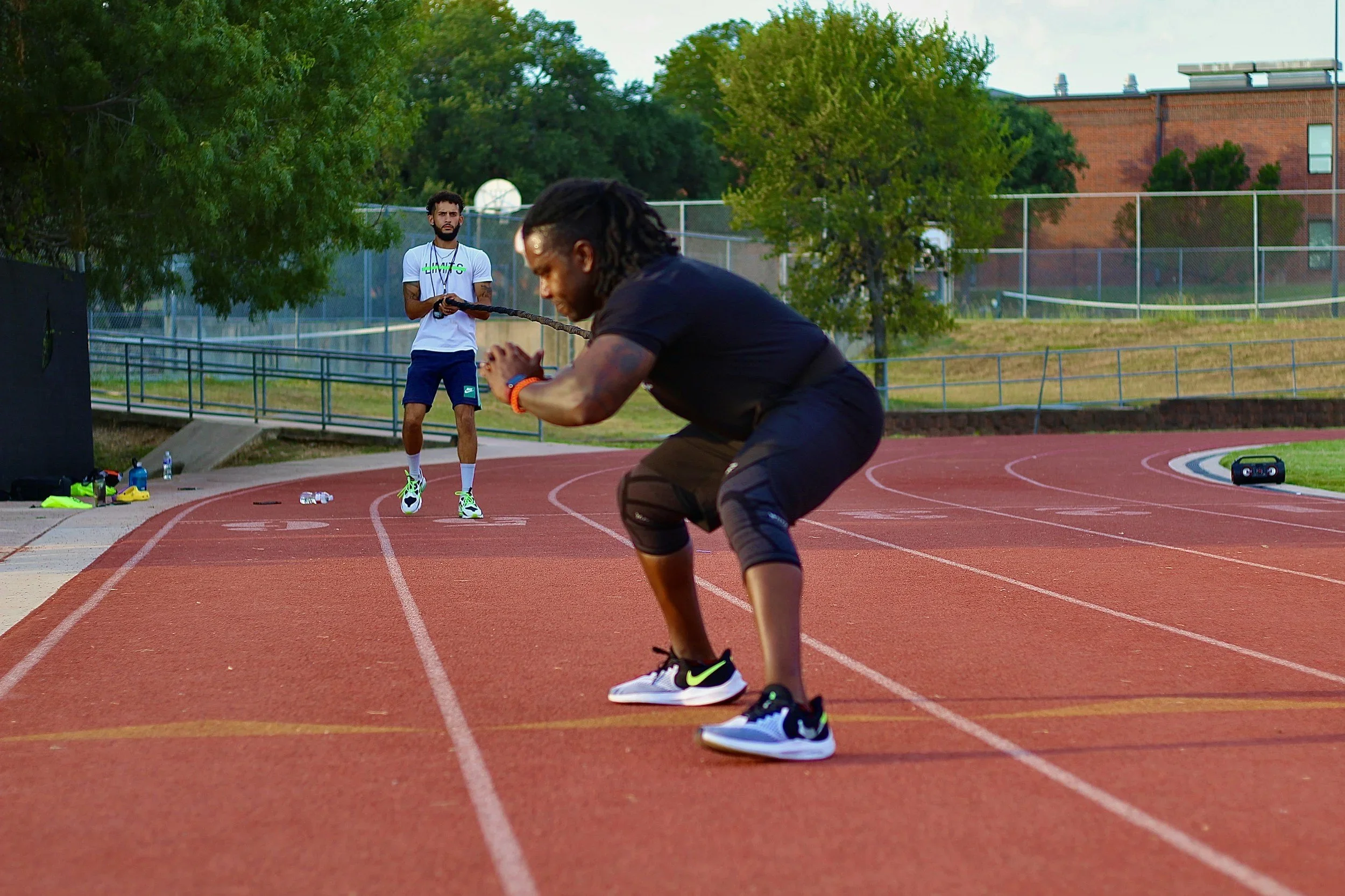 Two men on a red running track in a park. One man is in a crouched position, looking down with his hands clasped, and the other is standing in the background holding a resistance band, watching him. There are trees, a fence, and a brick building behind them.