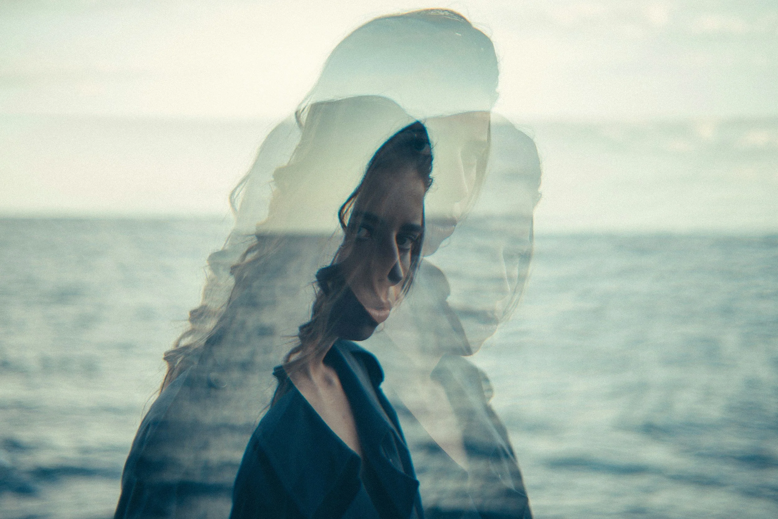 A woman standing near the ocean, with multiple overlayed semi-transparent images of her face creating a layered effect.