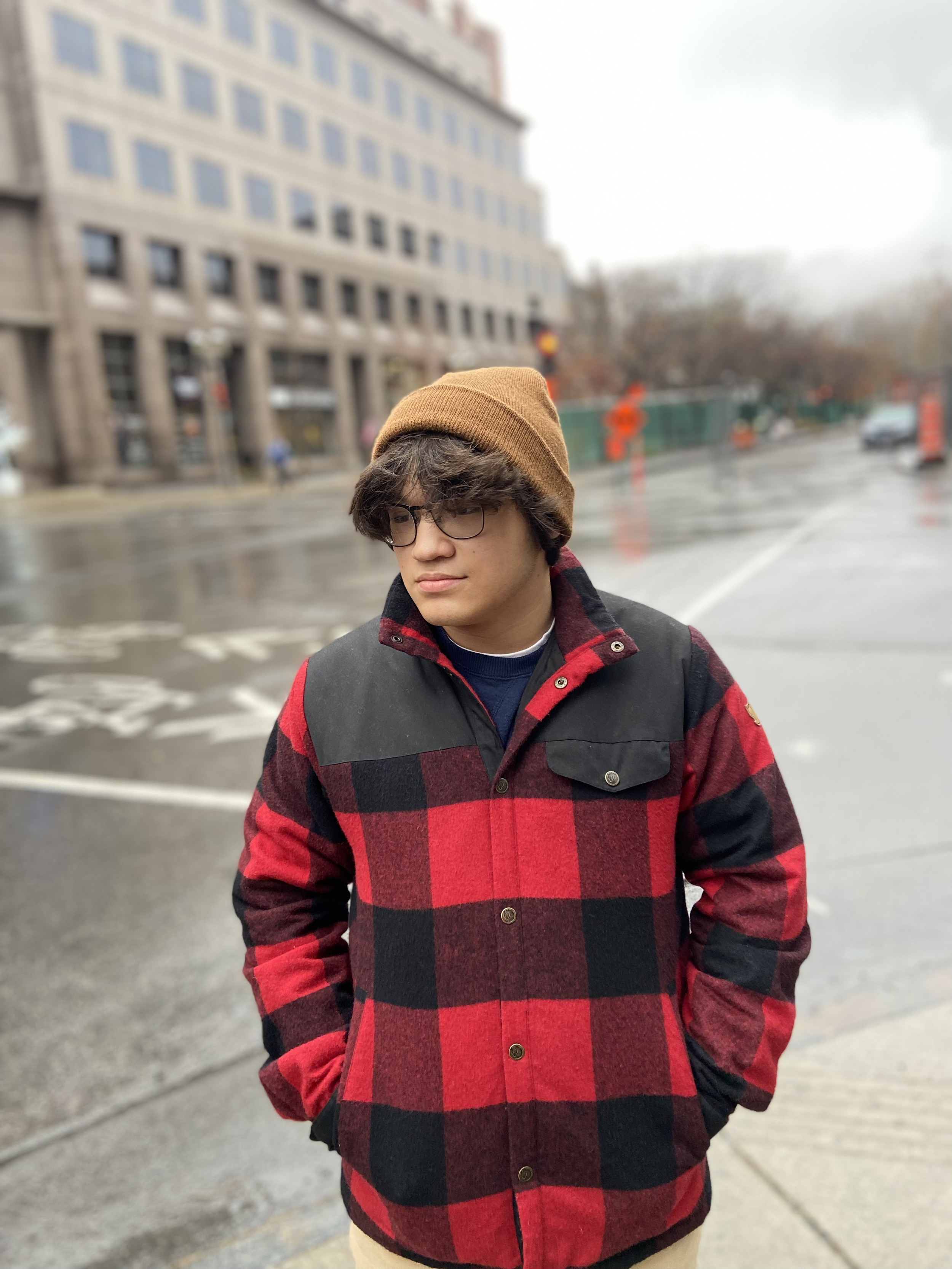 Young man with glasses, wearing a brown beanie and a red and black plaid jacket, standing on a rainy city street.