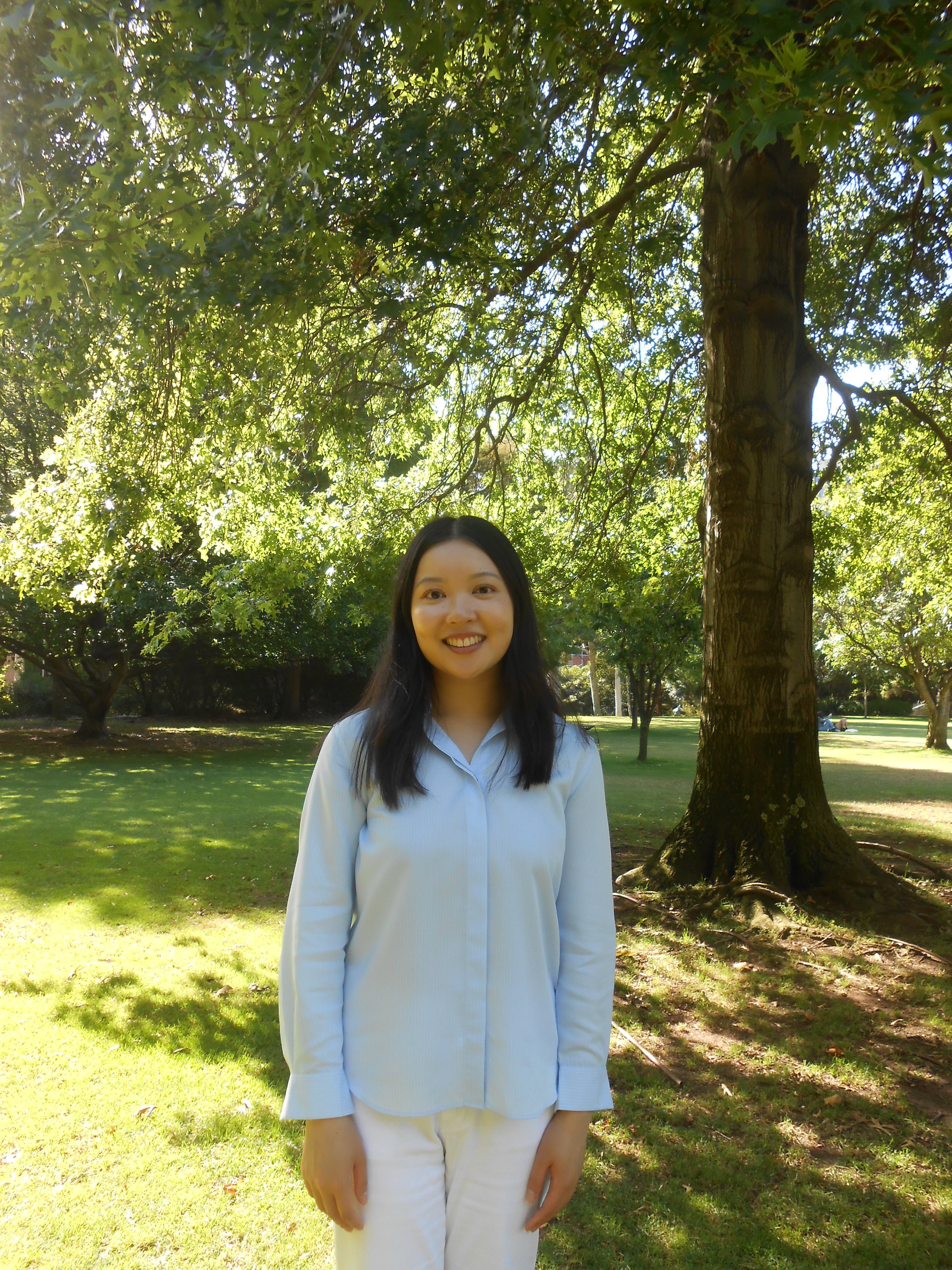 A young woman wearing a white shirt and white pants standing outdoors in a park with green grass and trees, smiling at the camera.
