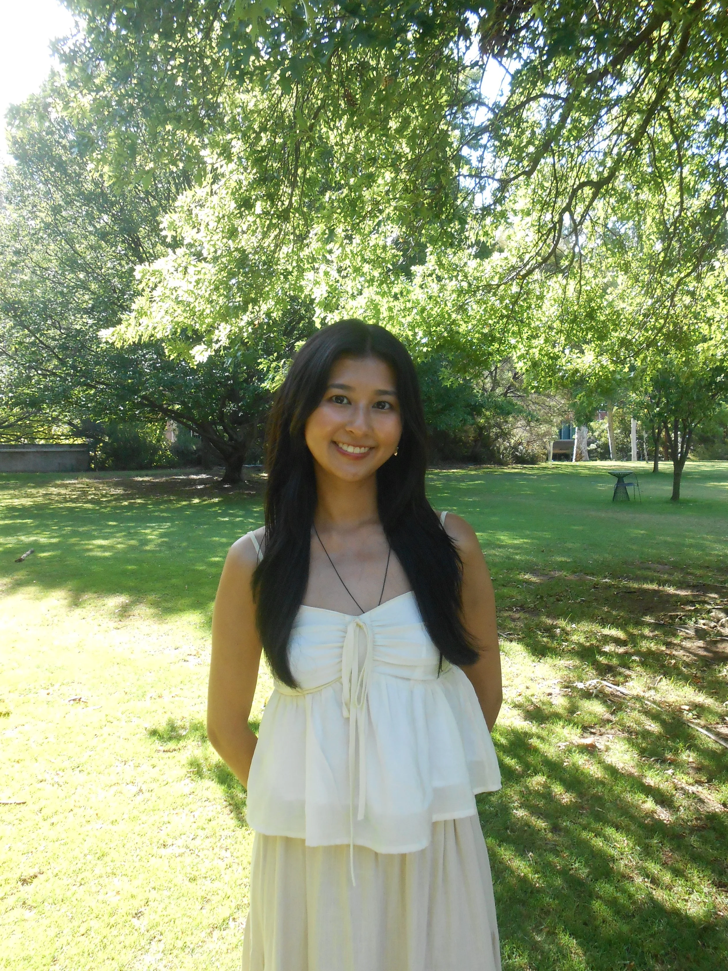A woman with long black hair smiling outdoors in a park with green trees and grass, wearing a white sleeveless dress.