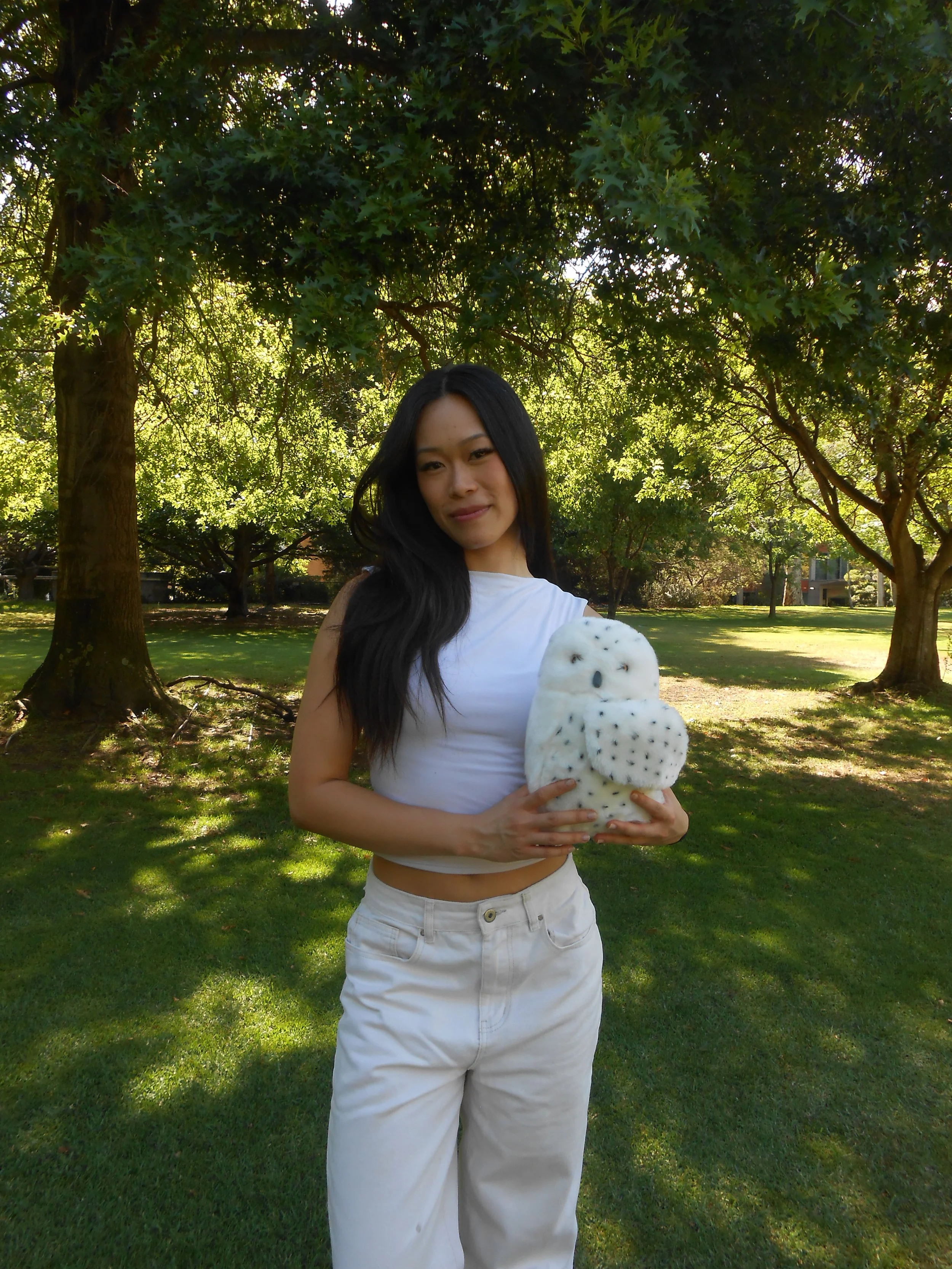 A woman with long black hair holding a plush owl toy outdoors in a park with green trees and grass.
