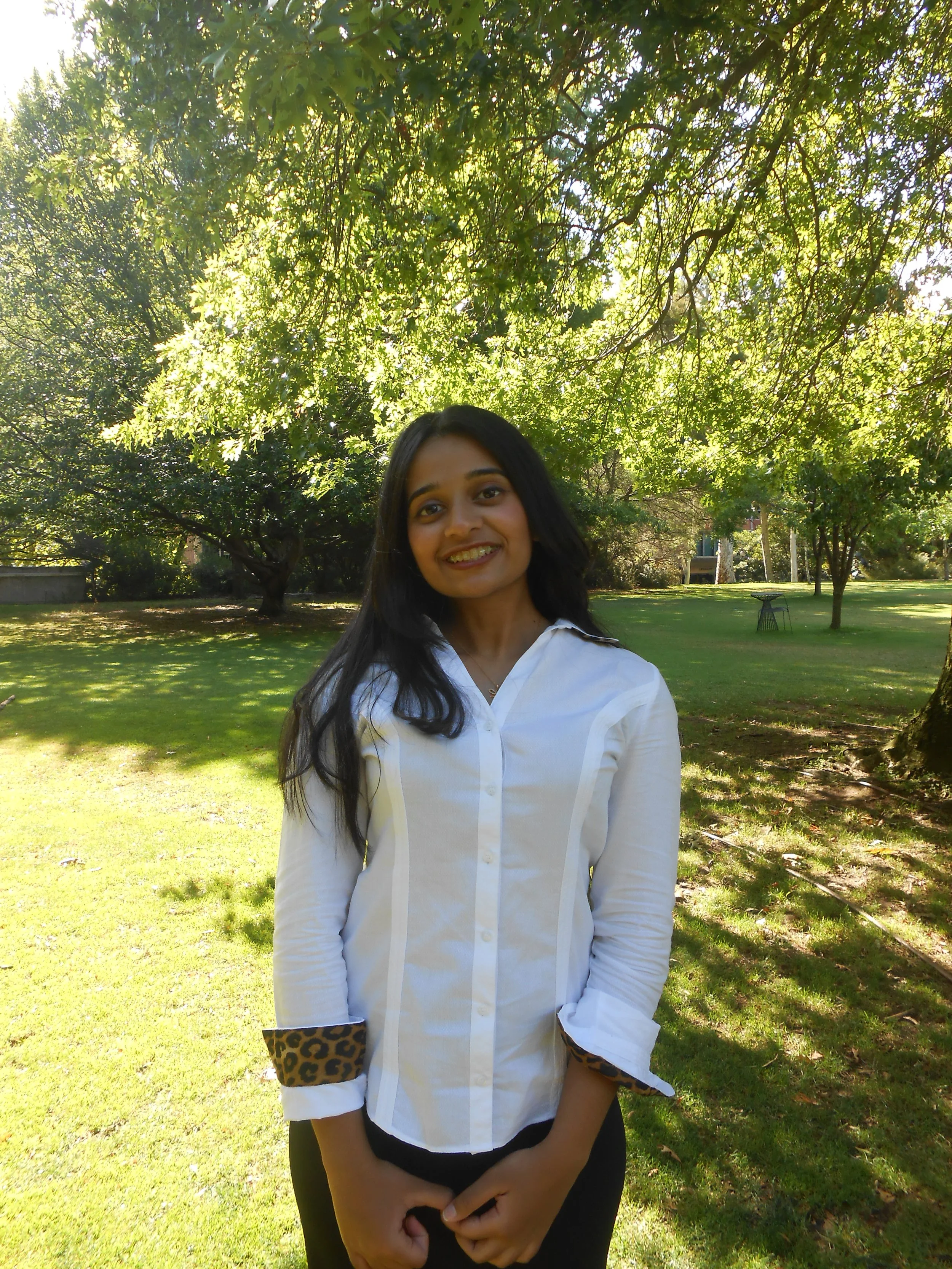A young woman with black hair and a white shirt with leopard print cuffs standing outdoors on a sunny day, with green trees and grass in the background.