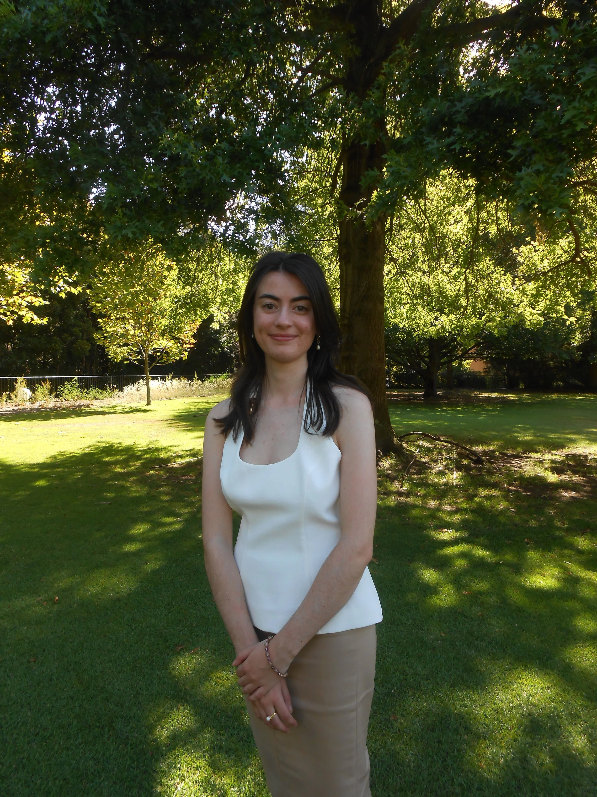 A woman in a white sleeveless top and beige skirt standing in a park with large trees and green grass, smiling at the camera.