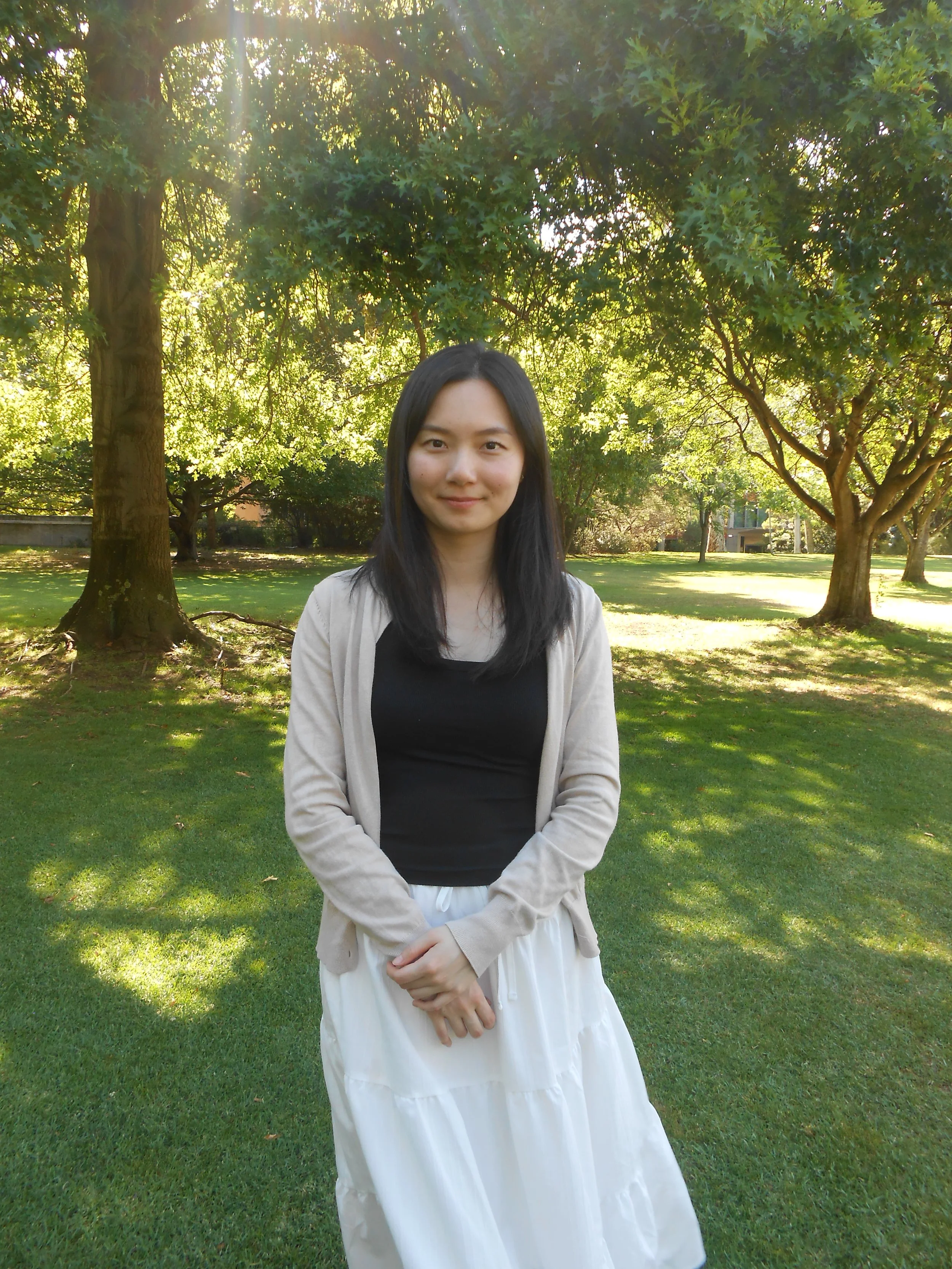 A young woman standing in a park with green grass and trees, sunlight filtering through the leaves.