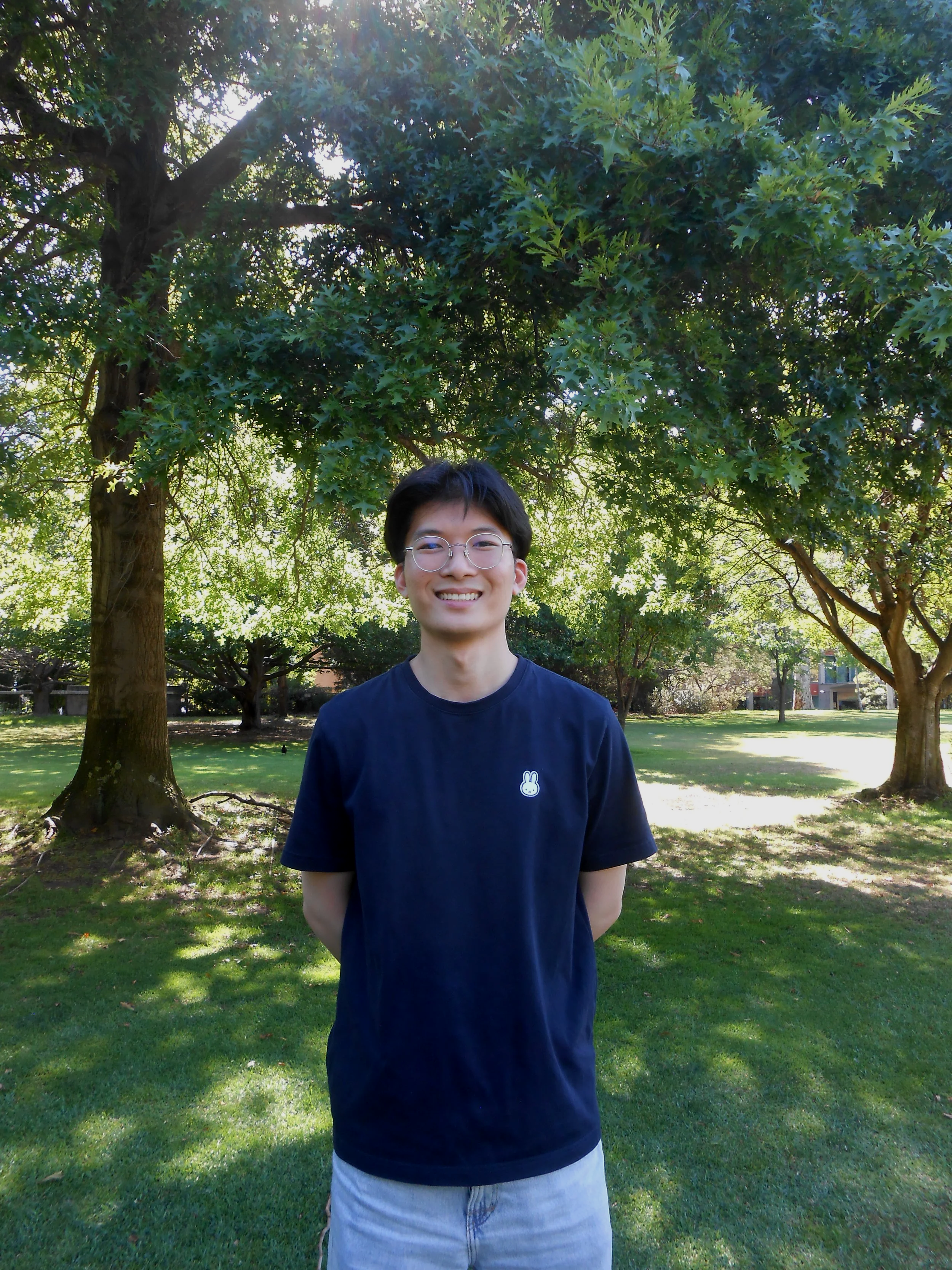 A young man with short black hair, wearing glasses, a navy T-shirt with a white bunny logo, and light-colored shorts, standing in a park with green grass and trees, smiling at the camera.