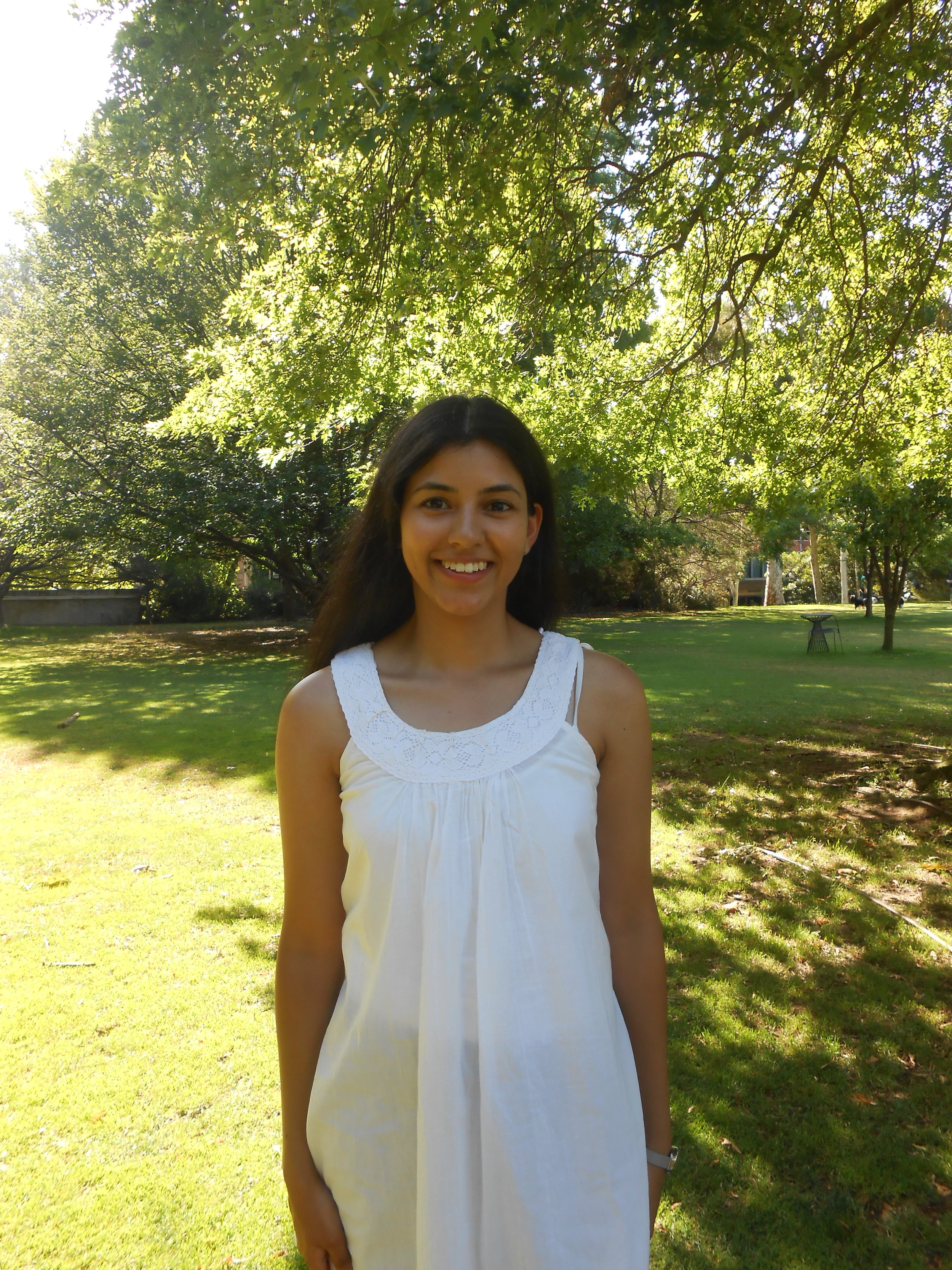 Young woman smiling in a white dress standing in a park with trees and grass.