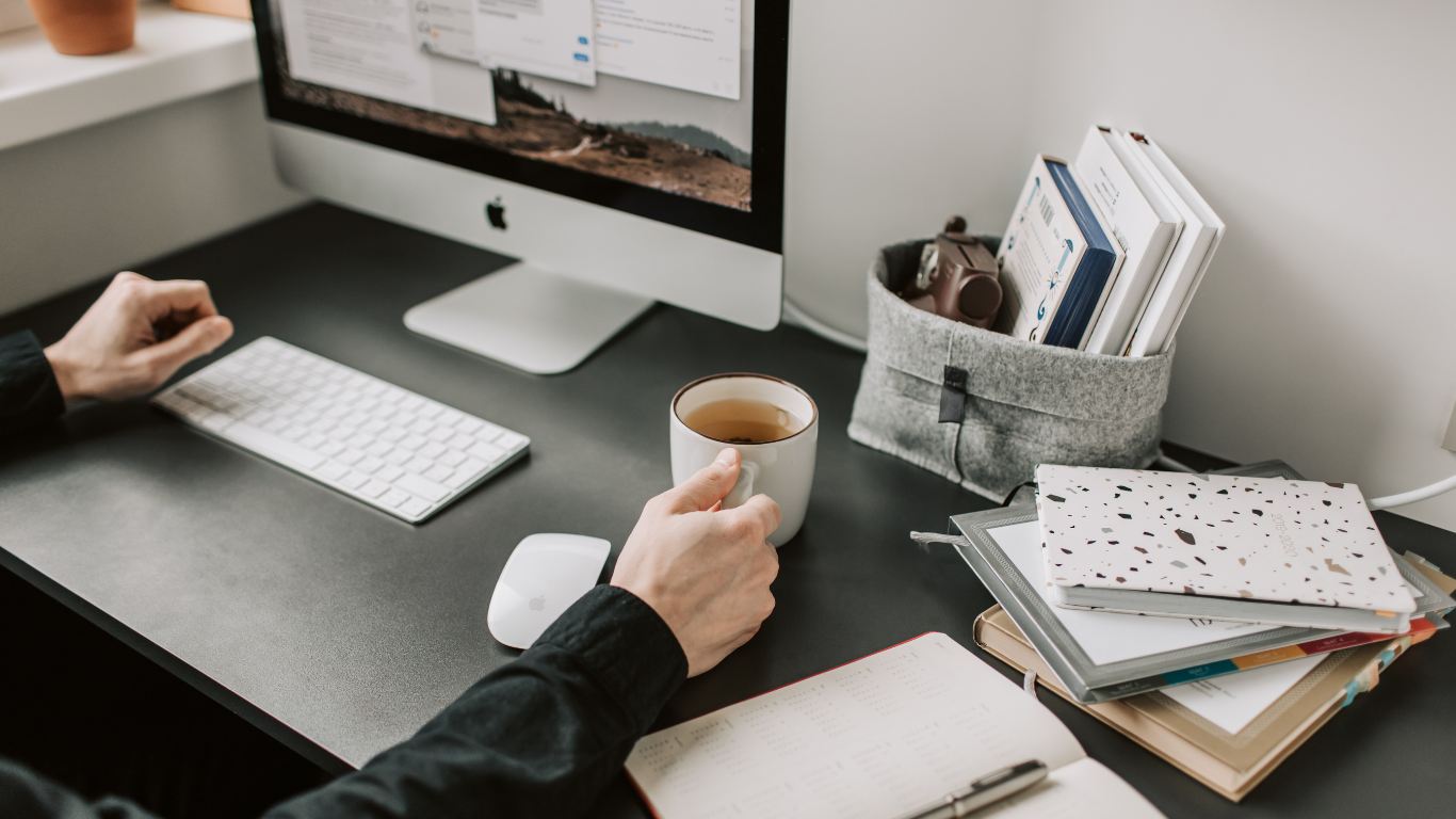 A person's hand holding a mug with tea on a black desk, with a desktop computer, a white keyboard, a white mouse, a stack of notebooks, and a gray fabric organizer filled with books and a camera.