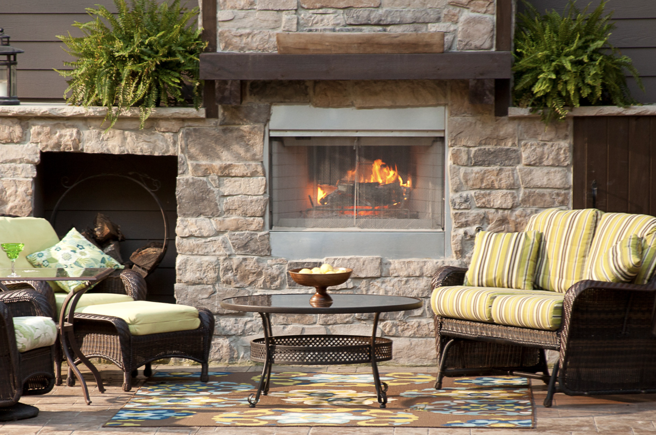Outdoor patio with wicker furniture, cushions, a stone fireplace with fire, and decorative plants.