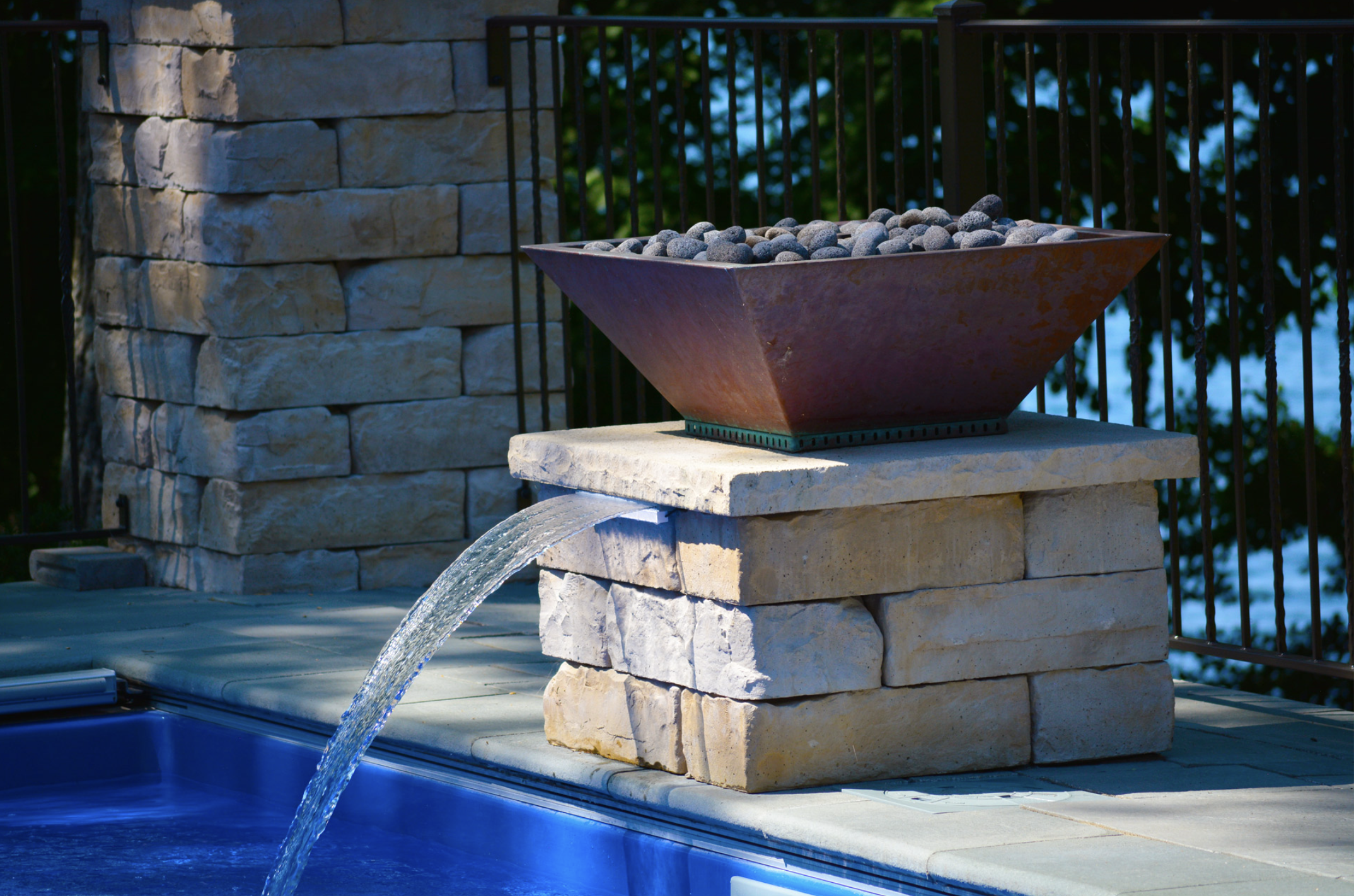 Water feature with a stone base and metal bowl of rocks, water flowing into a pool, surrounded by stone wall and railing.