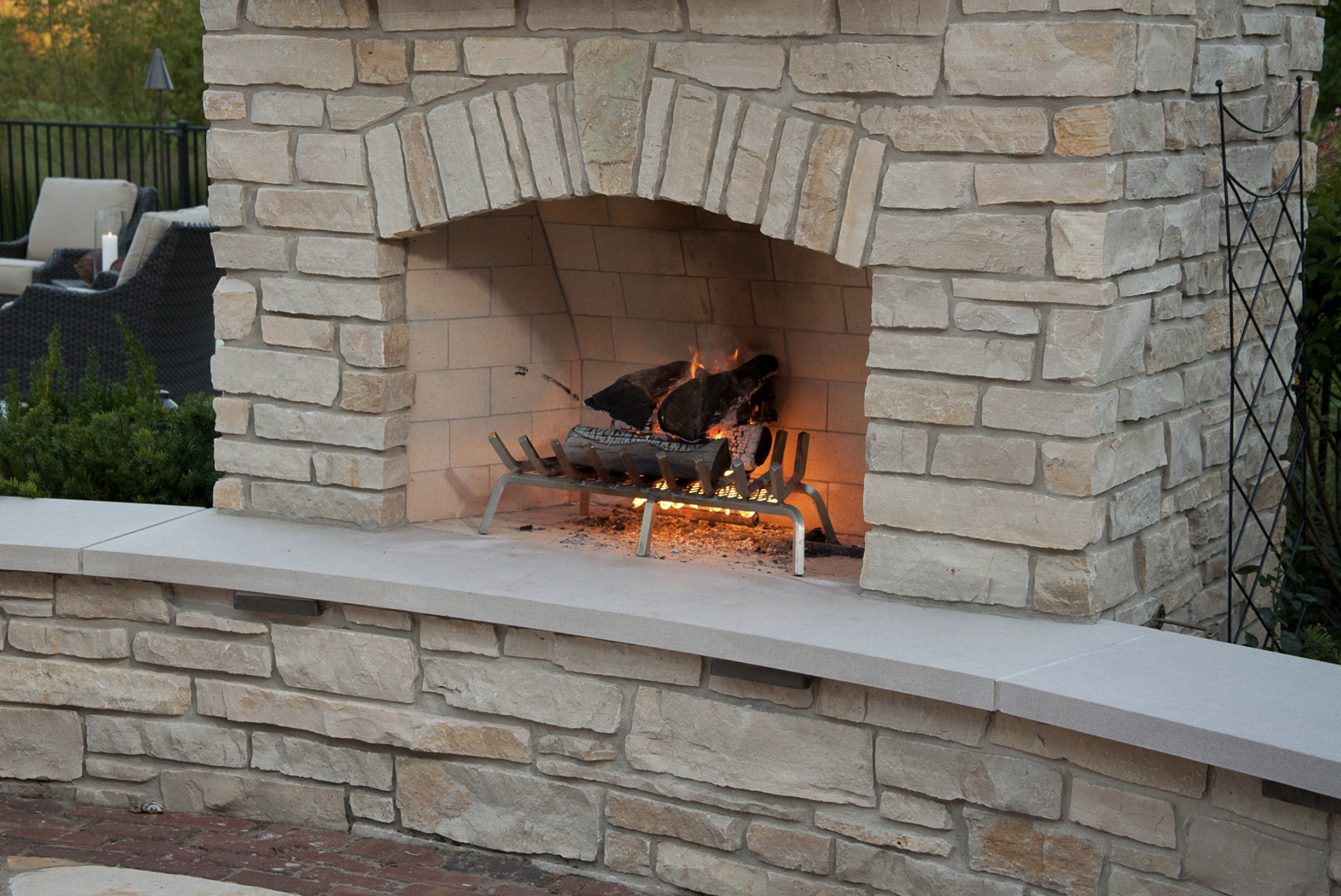 Outdoor stone fireplace with a lit fire and logs on a grate, surrounded by a brick and stone patio area.