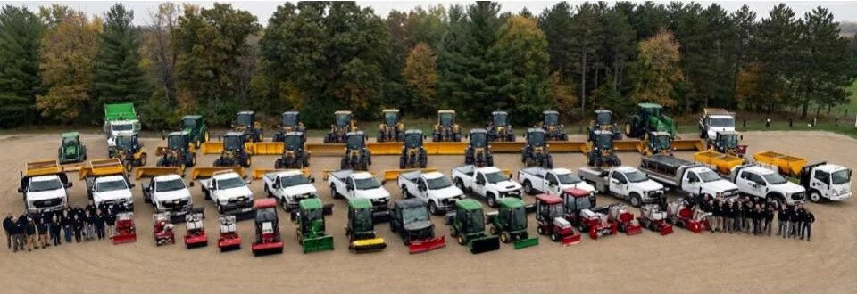Snow plow trucks and tractors with John Deere front loaders parked on a gravel lot with green fields and trees in the background.
