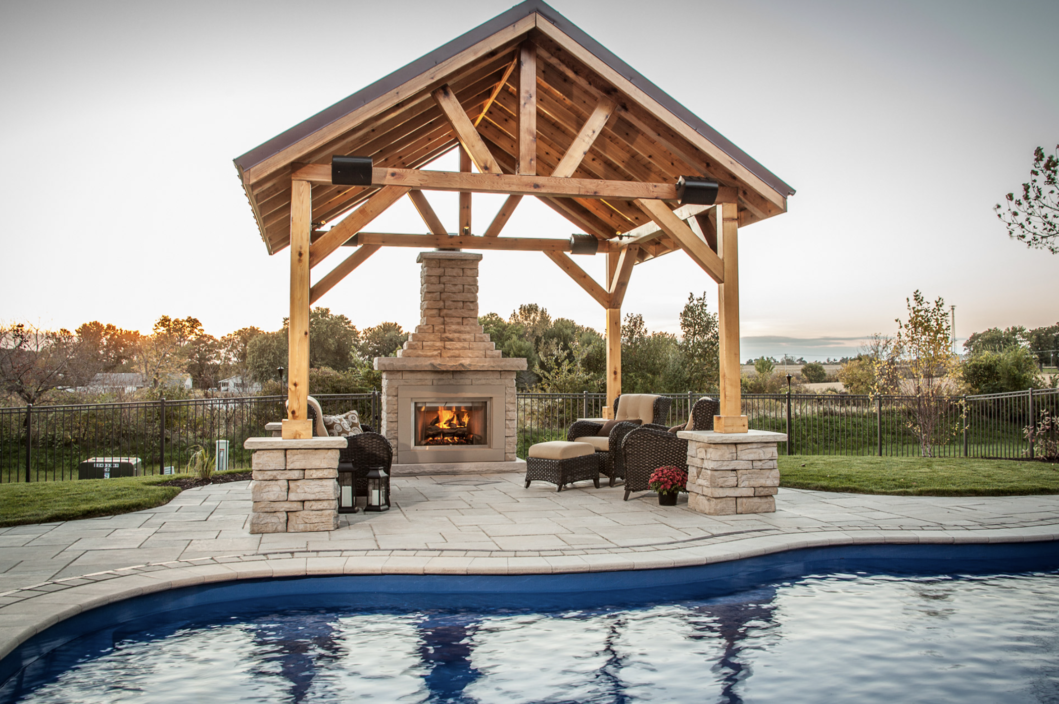 Outdoor patio with a wooden pergola, stone fireplace, wicker furniture, and a pool in the foreground.