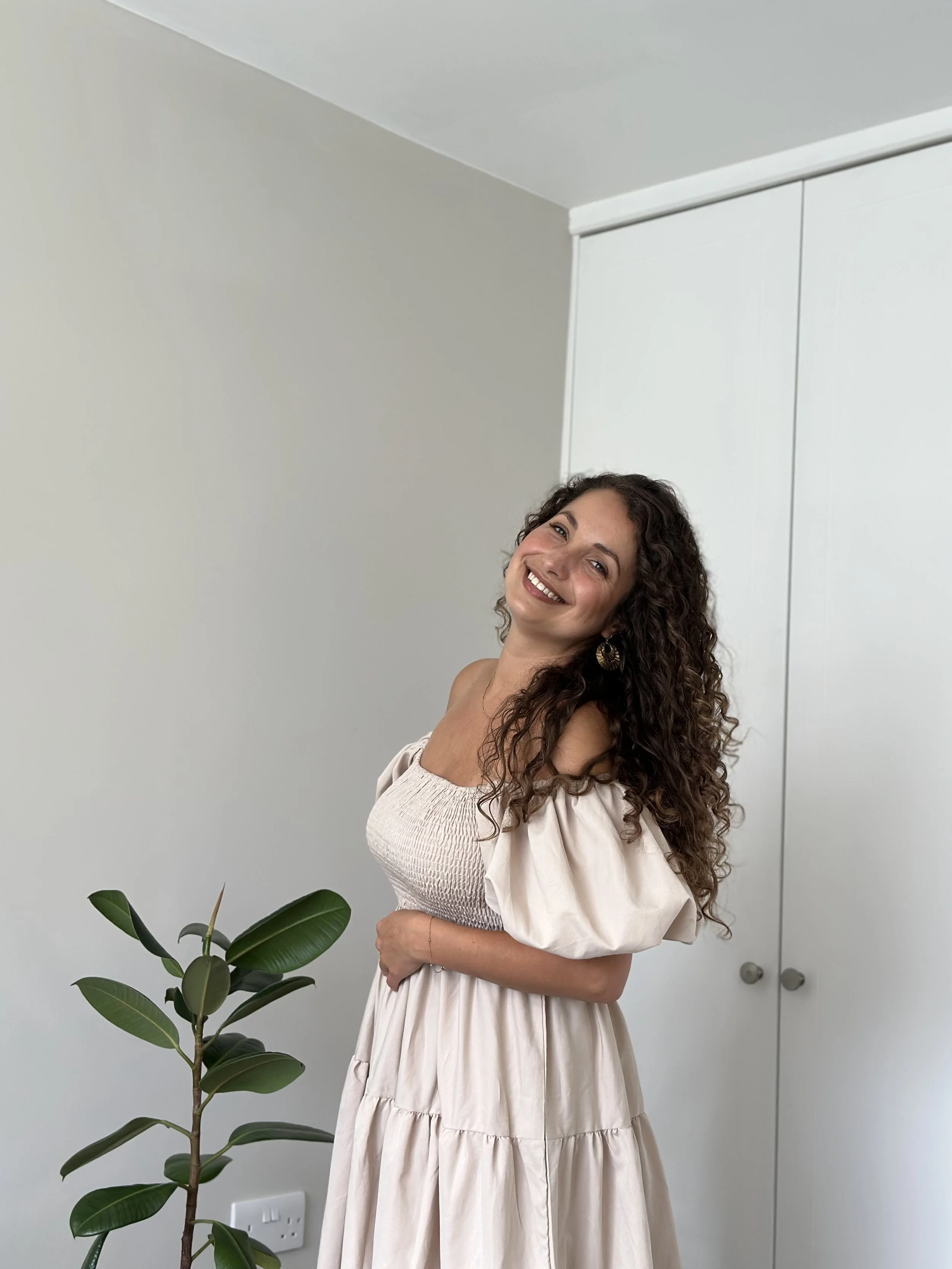 A smiling woman with curly hair wearing a beige off-shoulder dress standing indoors next to a green plant, with a white wall and closet in the background.