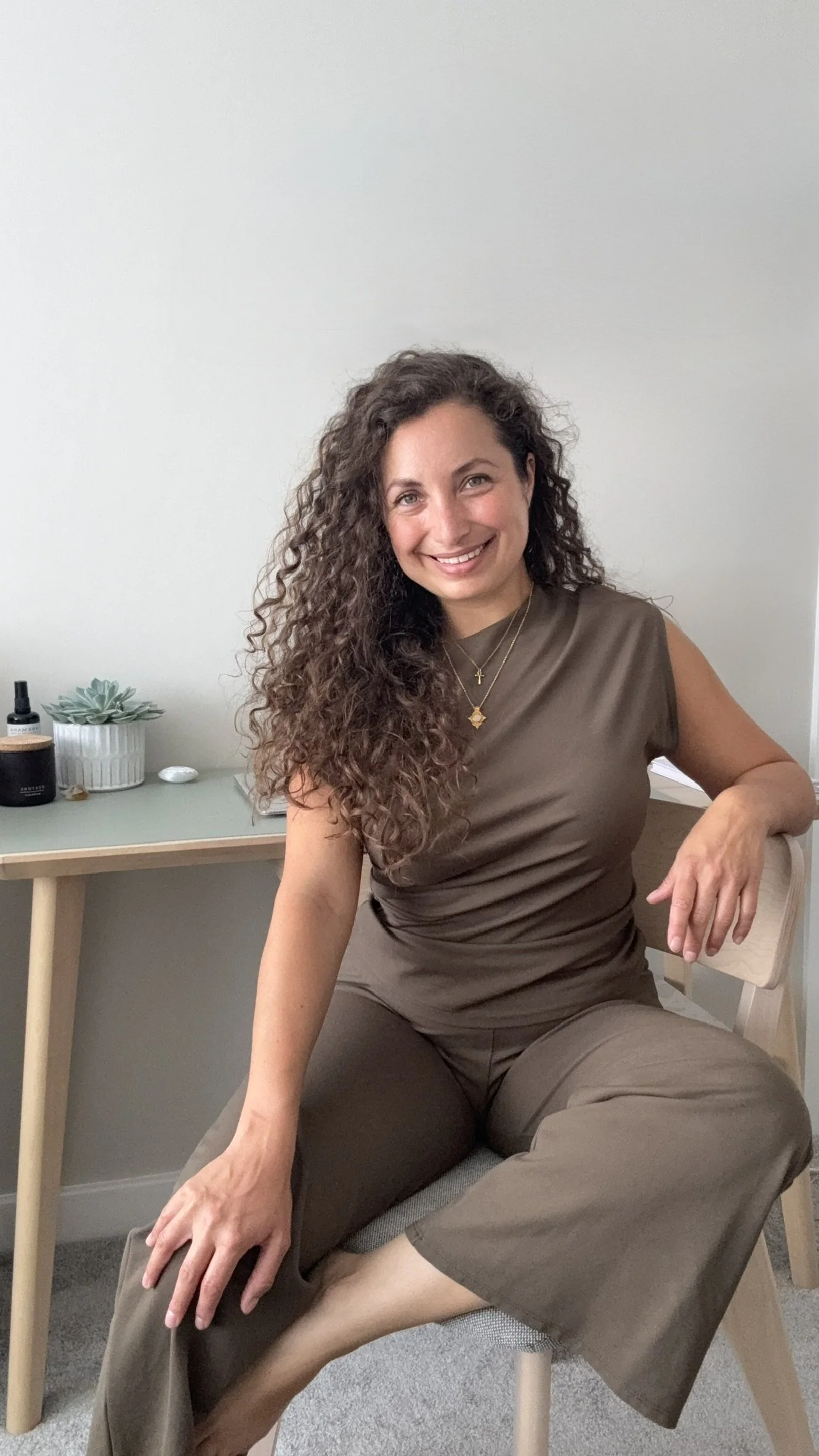 A woman with curly brown hair smiling, sitting on a beige chair in front of a light-colored wall, wearing a brown outfit and layered necklaces with cross and star charms, with a small table behind her holding a plant, perfume, and other items.